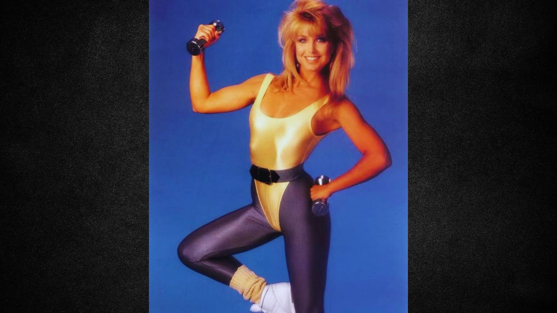Smiling woman in 80s workout attire holds dumbbells against a blue background. She wears a shiny leotard, leggings, and leg warmers, conveying energy.