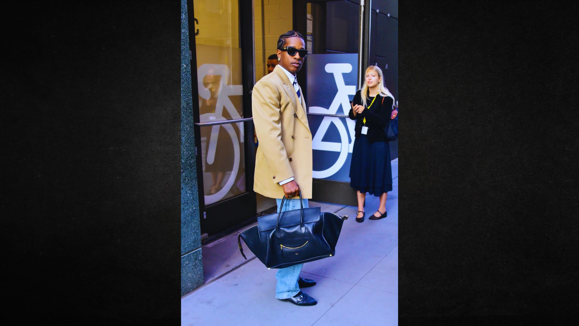 A man in a suit and sunglasses stands confidently, holding a stylish bag in his right hand.