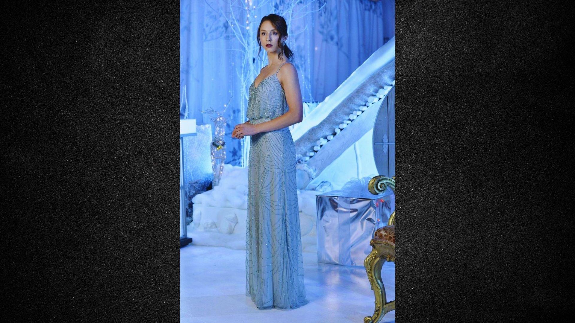 A woman in a blue dress stands in front of a mirror, gazing at her reflection with a thoughtful expression.