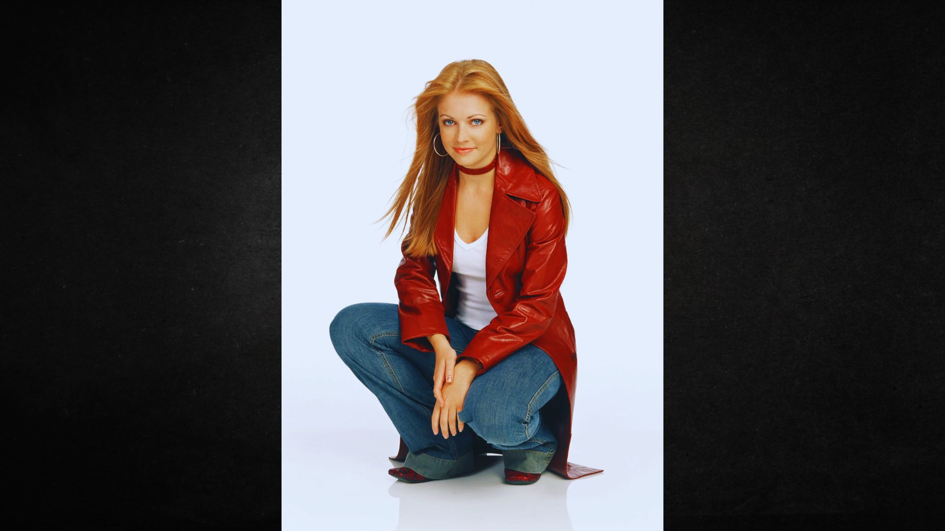 Young woman with long red hair crouches, wearing a red leather coat, white top, and blue jeans. She smiles softly against a light background.
