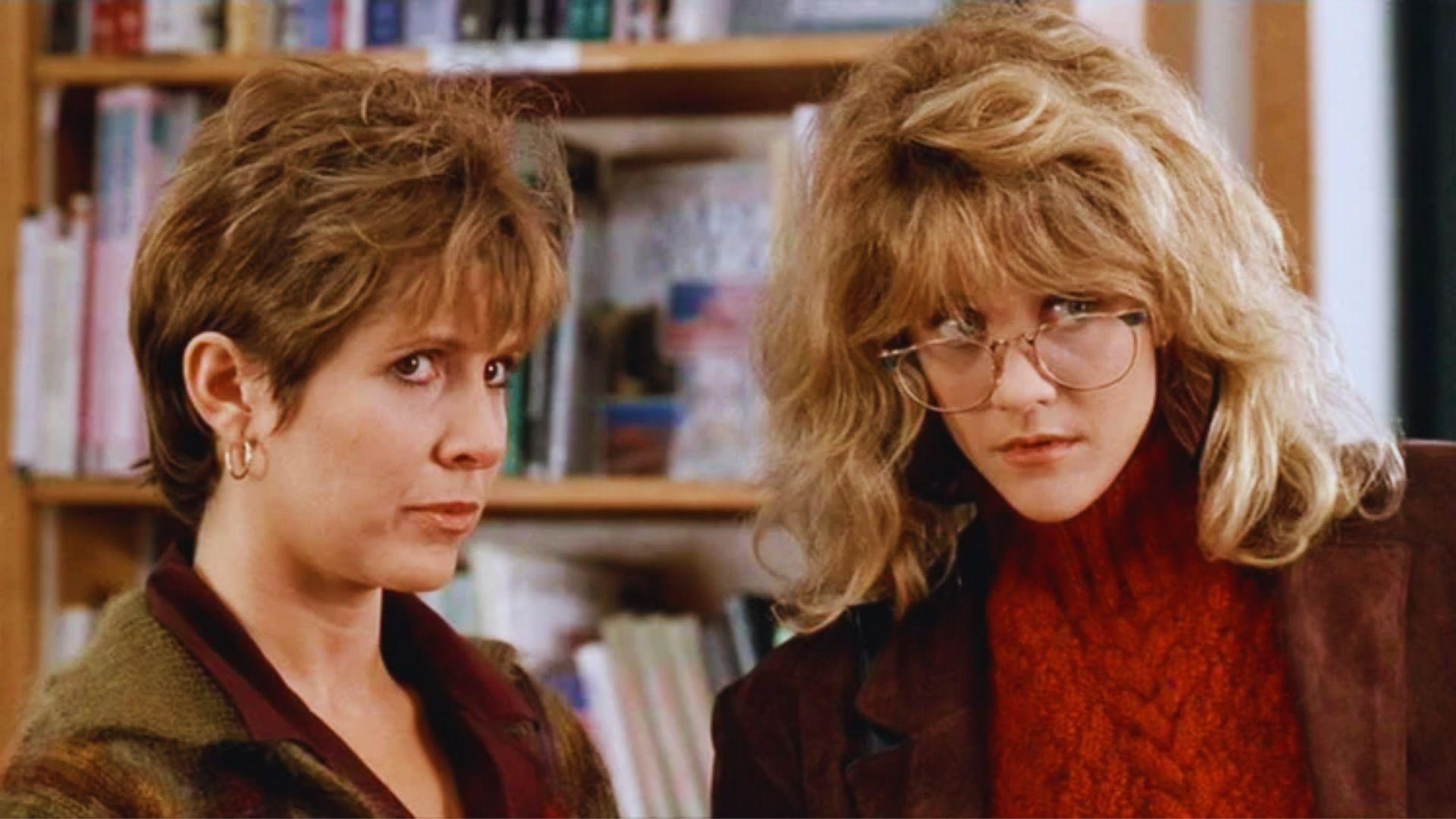 Two women with serious expressions stand indoors in front of a bookshelf. One has short hair and earrings; the other wears glasses and a knitted sweater.