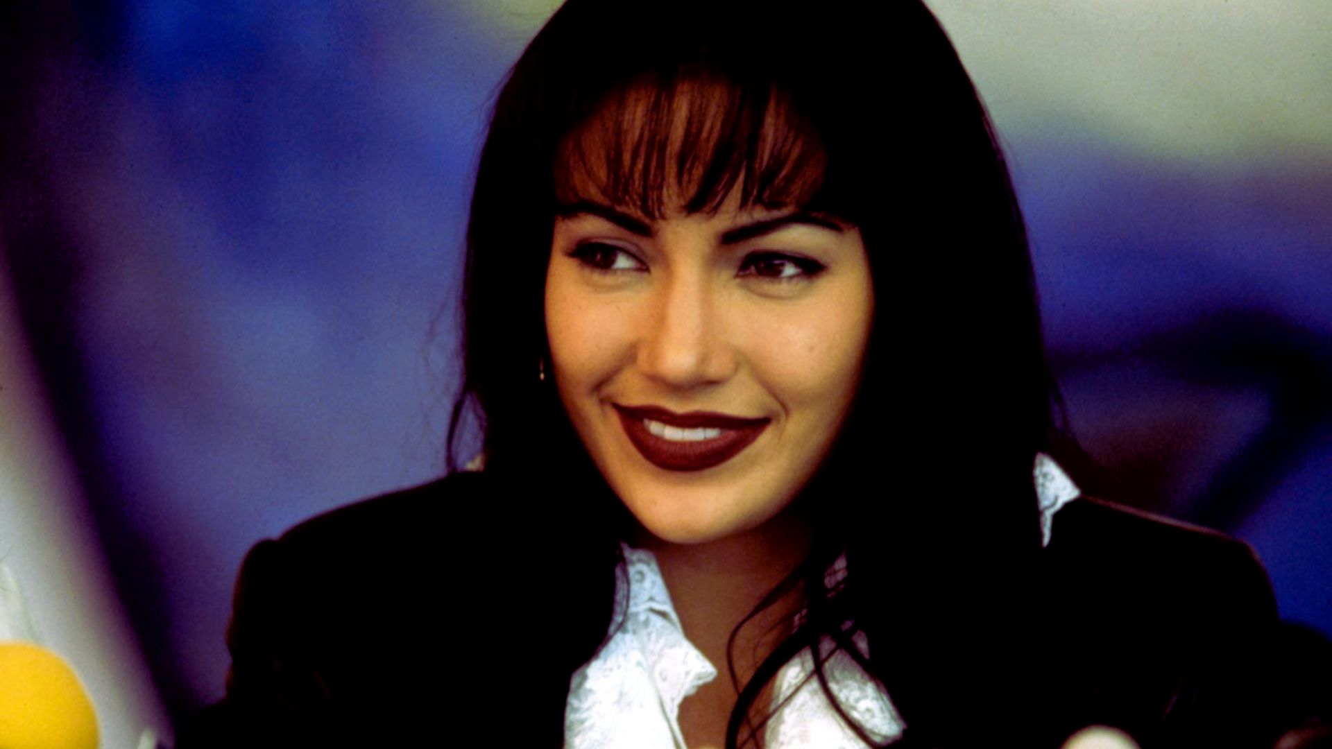 A woman with dark hair and bangs smiles warmly. She is wearing a white blouse and dark jacket. The background is softly blurred in blue hues.