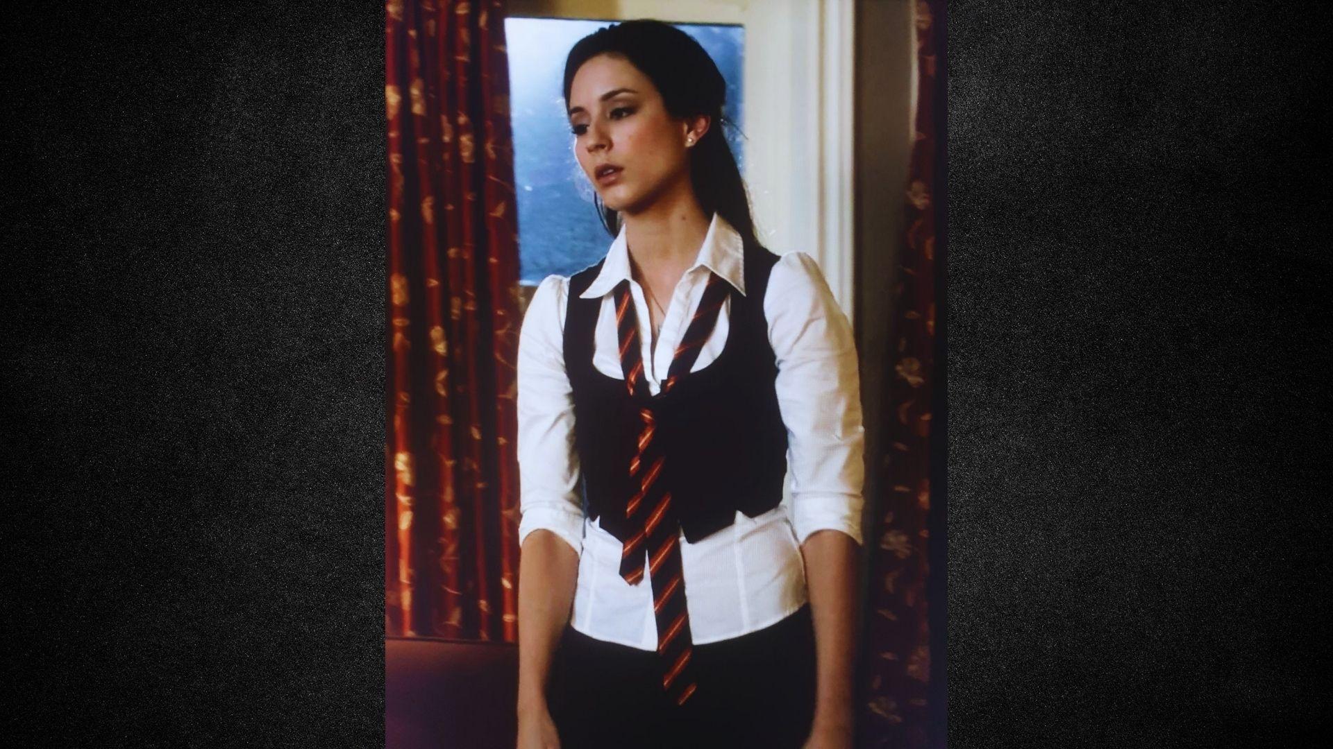 A woman wearing a tie and vest stands confidently in front of a large window, with natural light illuminating the scene.