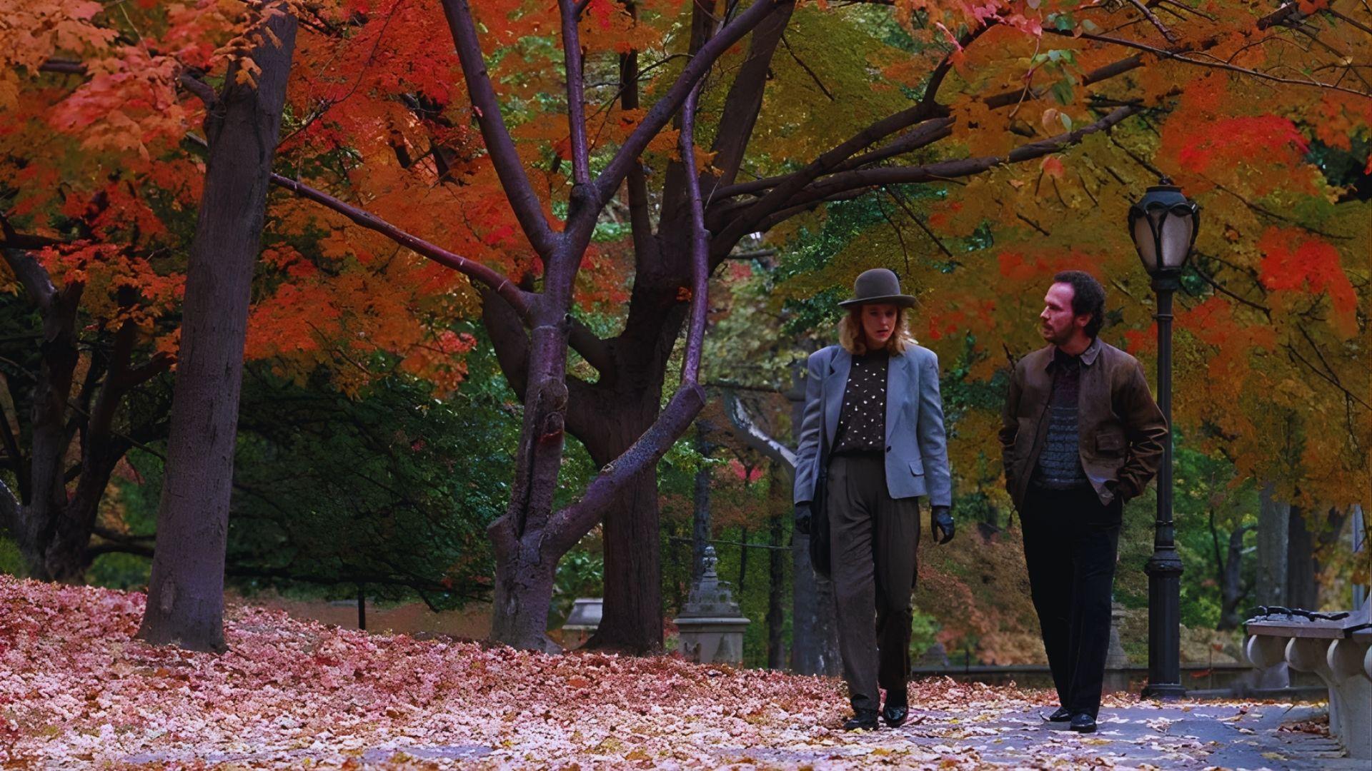 A man and woman walk through a park with vibrant autumn leaves in red, orange, and yellow hues around them, creating a warm, nostalgic atmosphere.