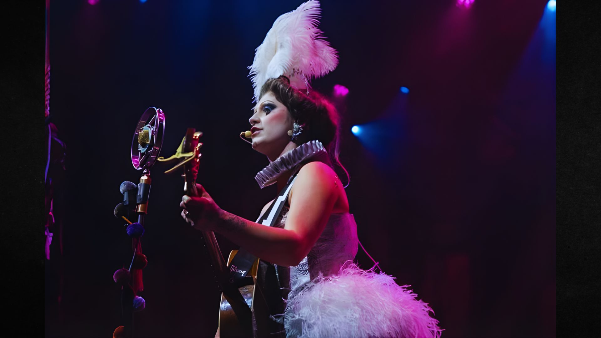 Performer in feathered costume and white headpiece sings into a retro microphone while playing guitar on a dimly lit stage. Dramatic and theatrical ambiance.