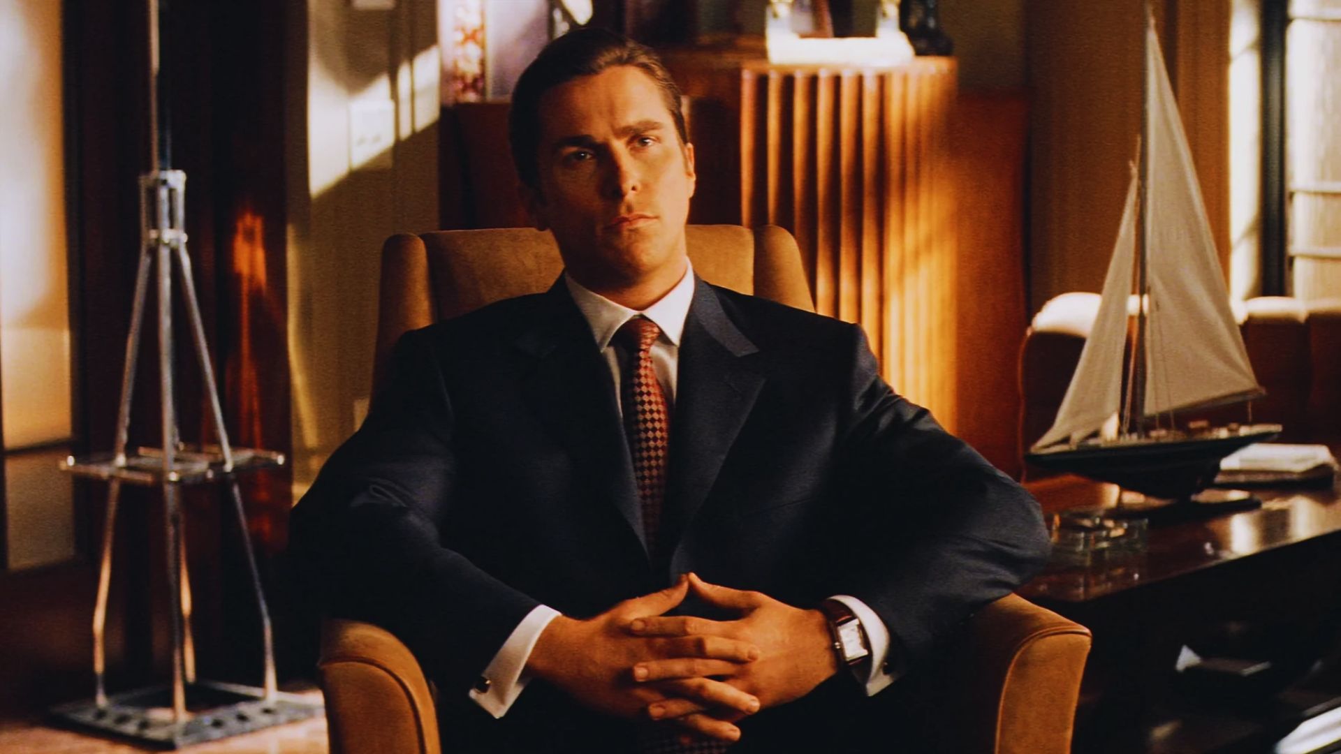A man in a suit sits confidently in a leather armchair in an elegant office. Behind him is a wooden shelf and a decorative sailboat, conveying sophistication.