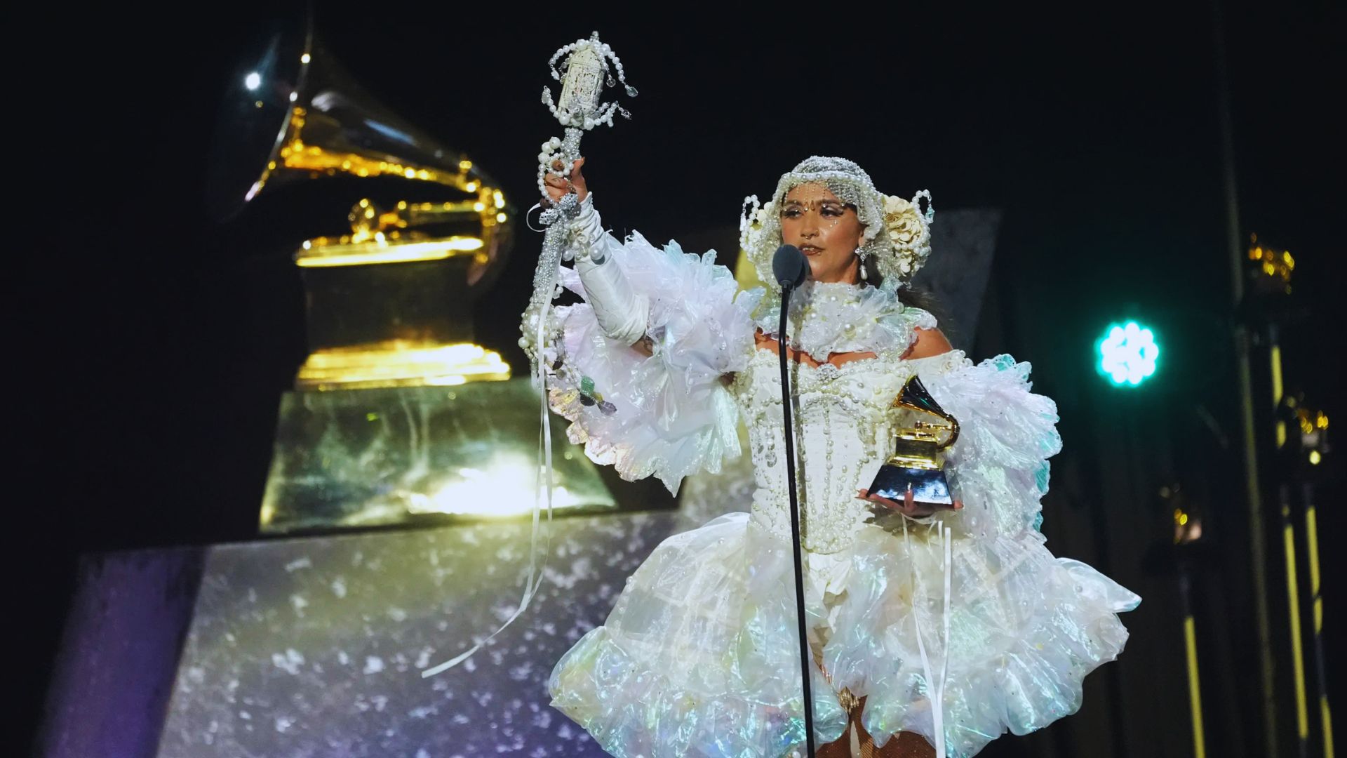 A performer in a white, ruffled, iridescent outfit holds a Grammy award while raising a bejeweled staff. A large Grammy statue is visible in the background.