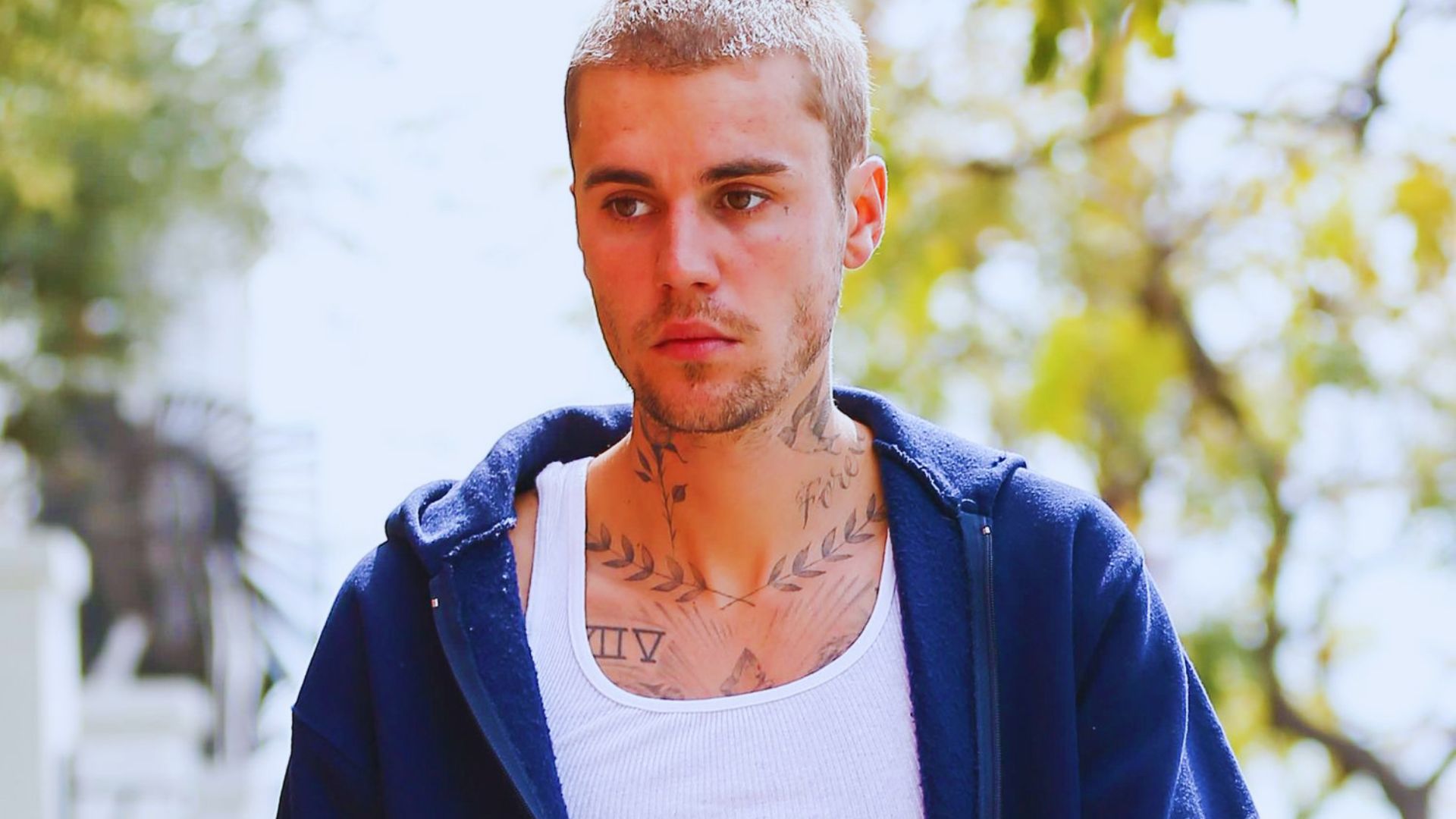 A tattooed man stands outside, showcasing intricate designs on his chest against a natural background.