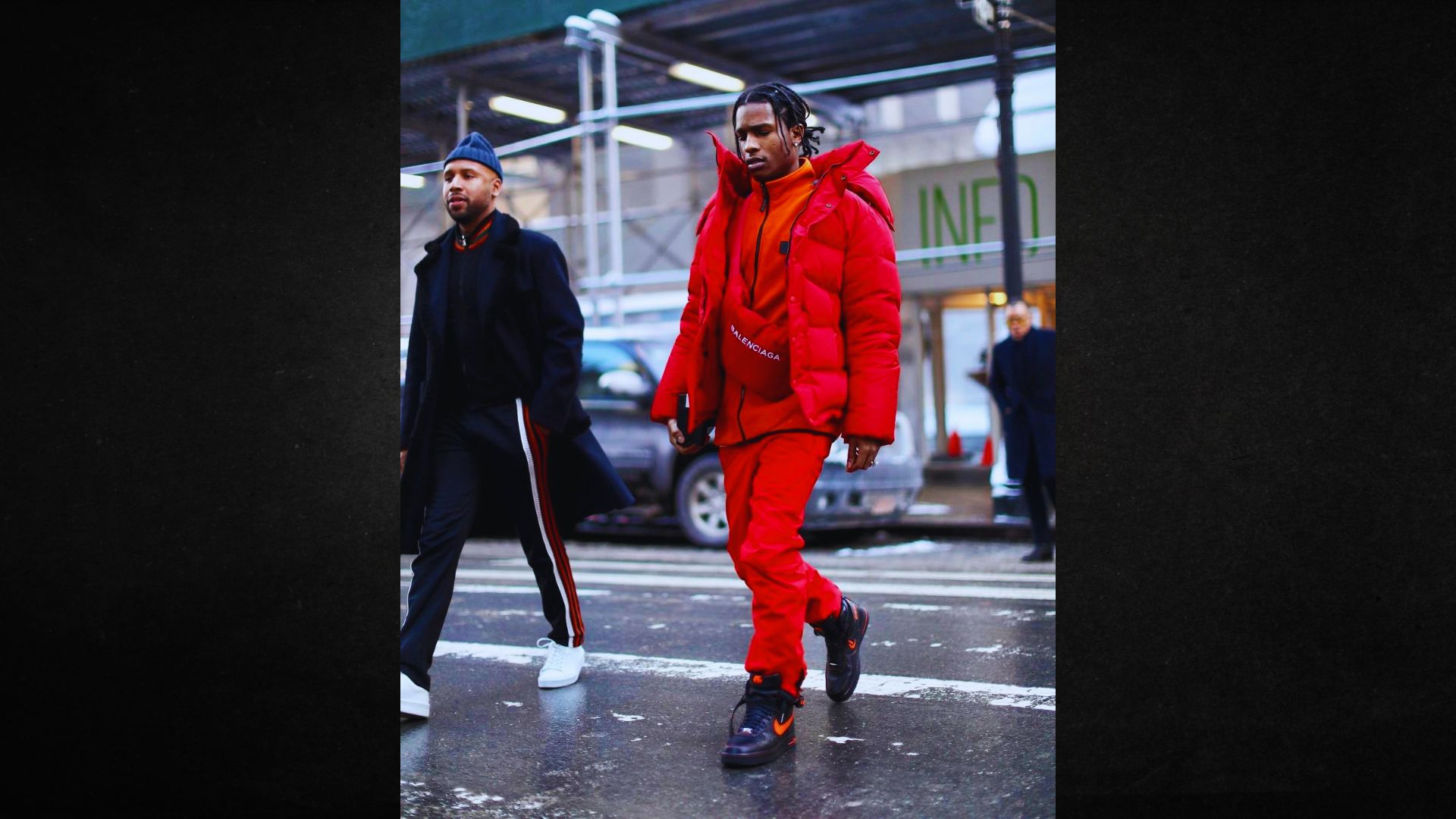 Two men walk on a city street. One wears an all-red outfit with a puffer jacket, exuding confidence, while the other is in a black coat and white sneakers. Urban setting.