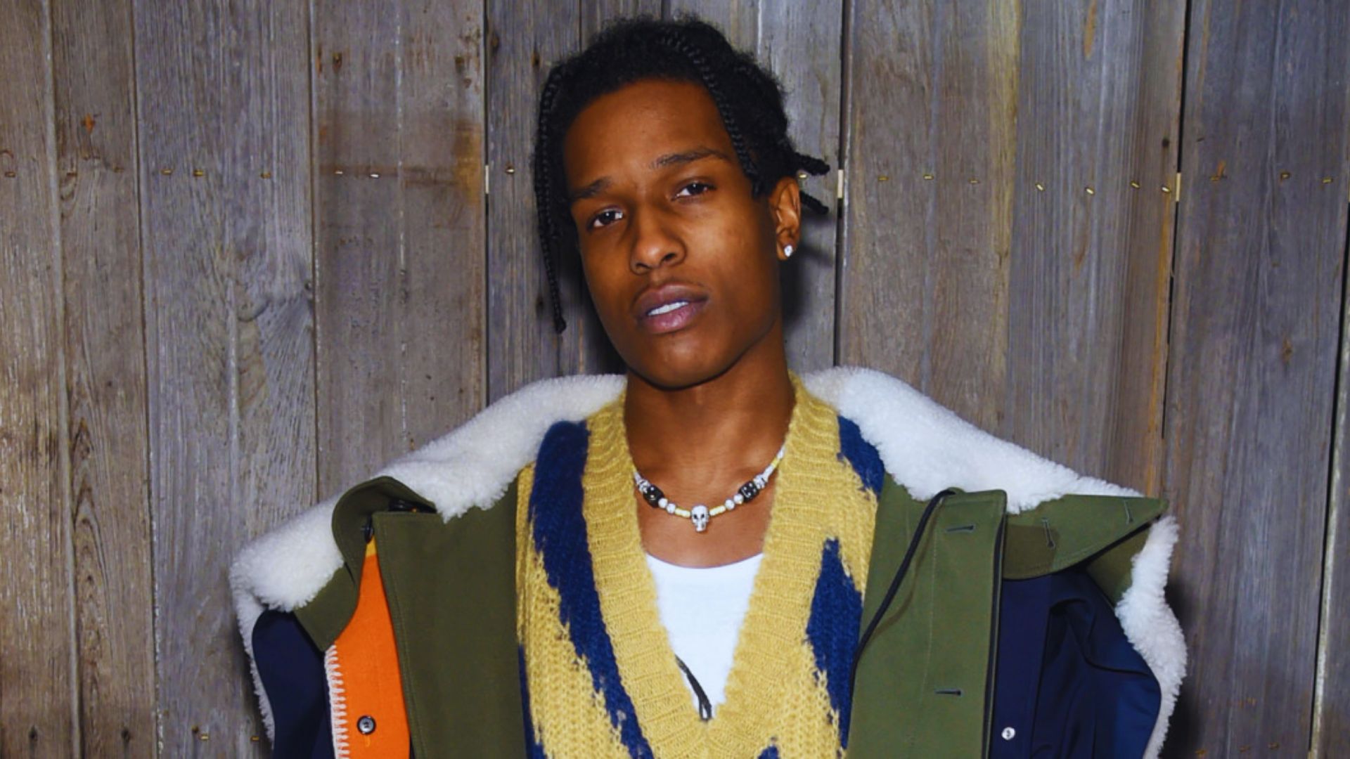 A person stands against a wooden fence, wearing a green and orange jacket with a cozy yellow and blue sweater. They convey a relaxed, confident vibe.