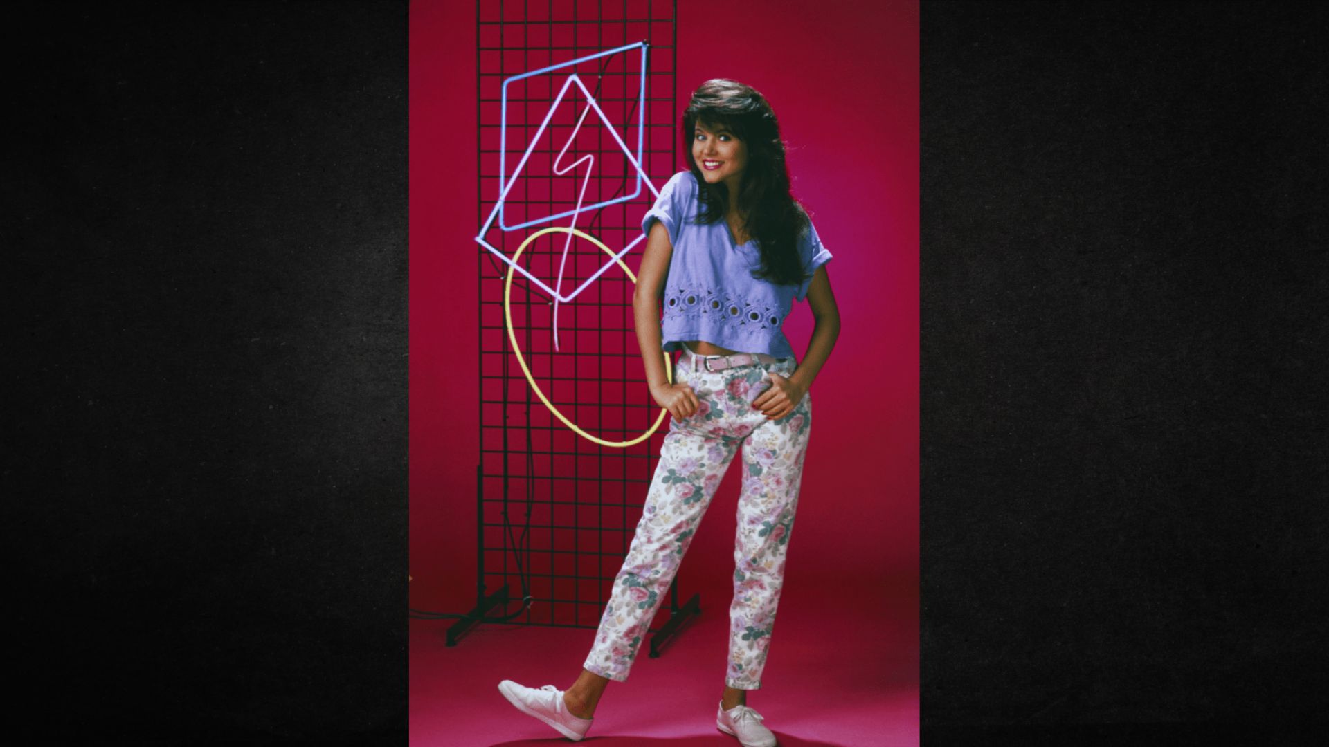 Teen girl in a blue top and floral pants stands against a vibrant pink backdrop. Neon geometric shapes add a retro 1980s vibe. Bright and stylish.