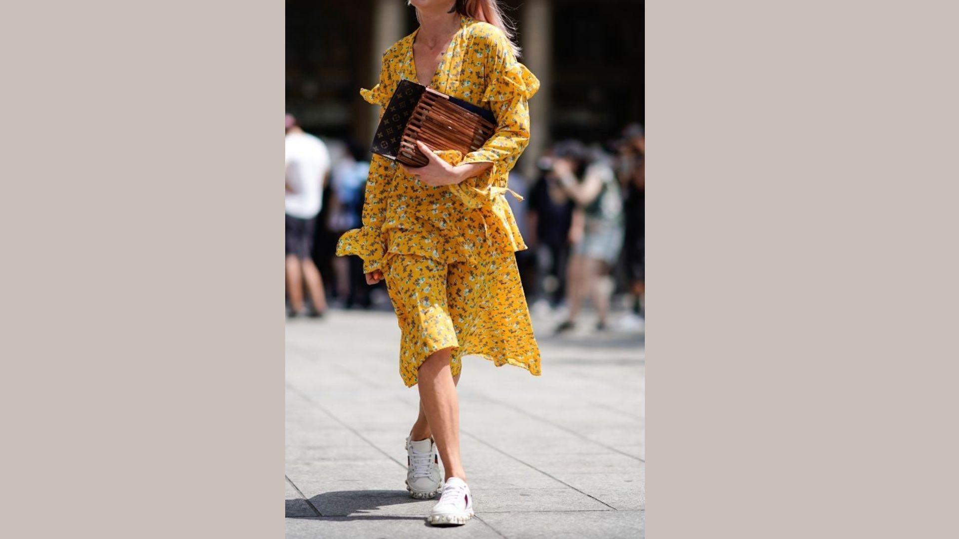 A woman wearing a yellow dress and white sneakers stands confidently, showcasing a vibrant and casual style.