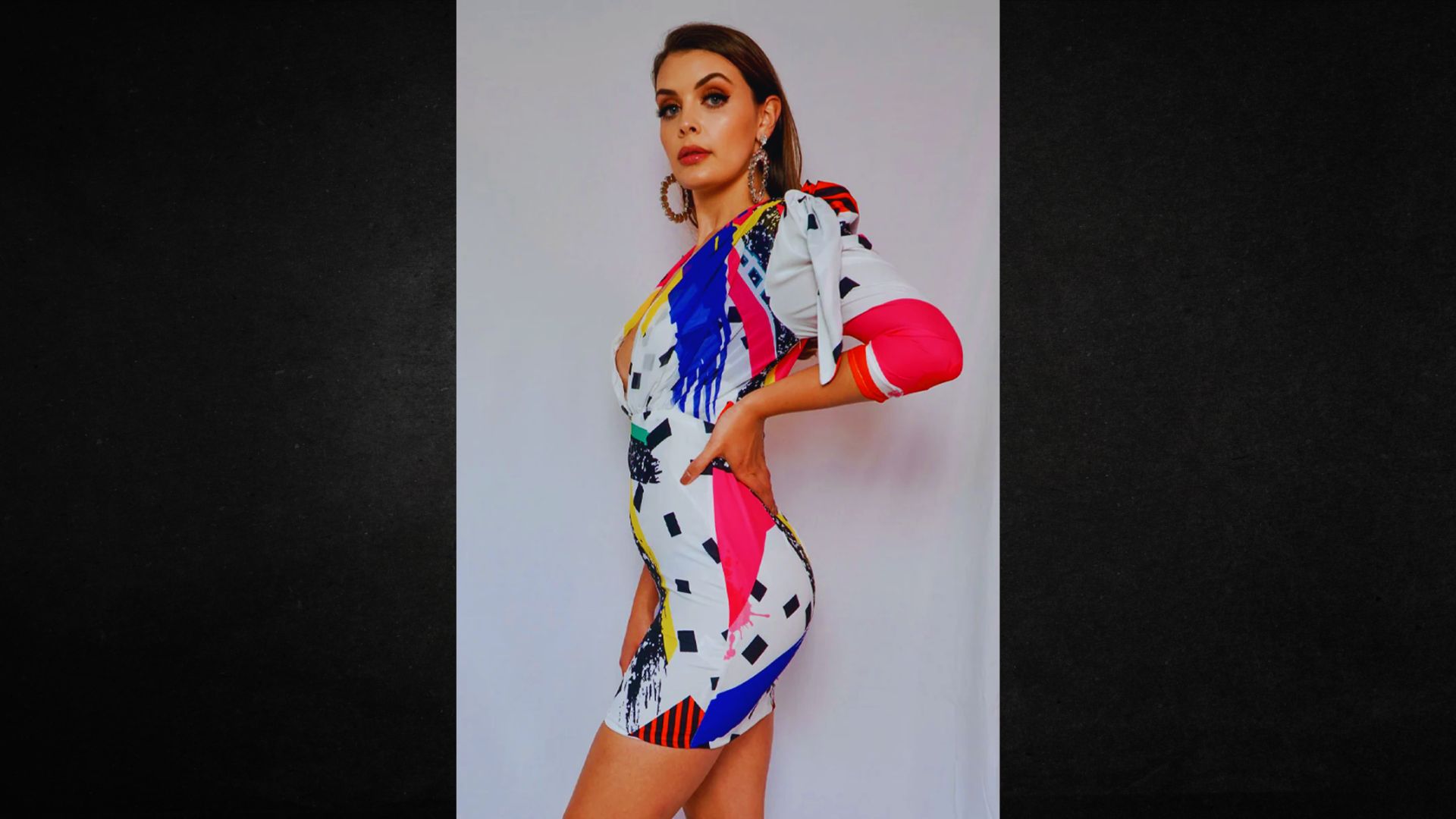 A woman poses confidently in a vibrant, abstract-patterned dress with bold colors like pink, blue, and yellow. Her expression is poised and stylish.