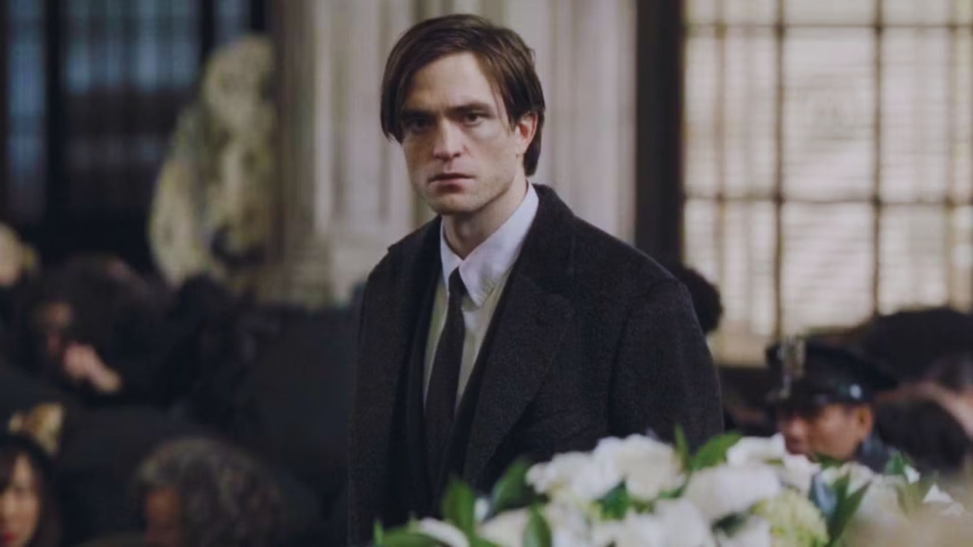 A man in a suit stands solemnly in a dimly lit room, surrounded by people and white flowers. The atmosphere conveys a sense of mourning and introspection.