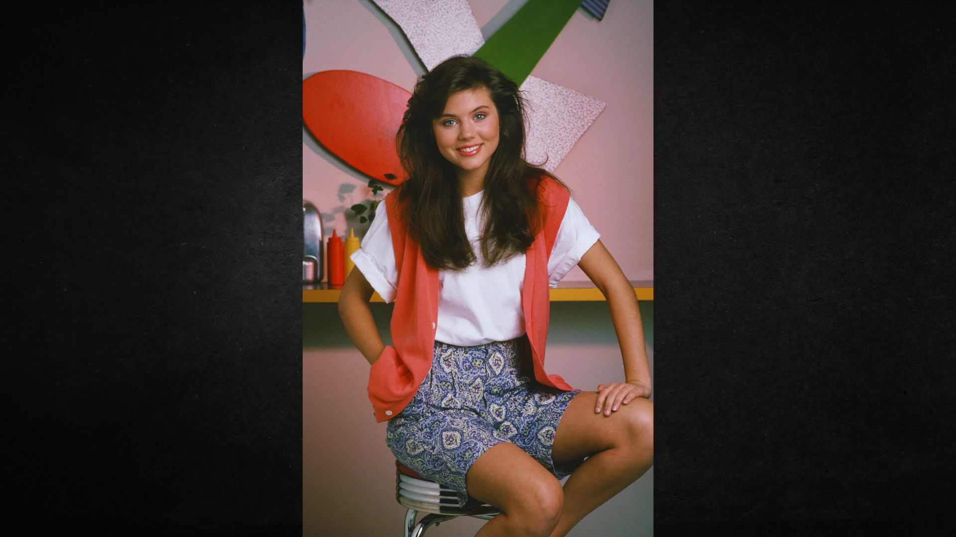 Young woman with long brunette hair sits on a stool, smiling. She wears a white shirt, red vest, and patterned shorts. Background has colorful abstract shapes.