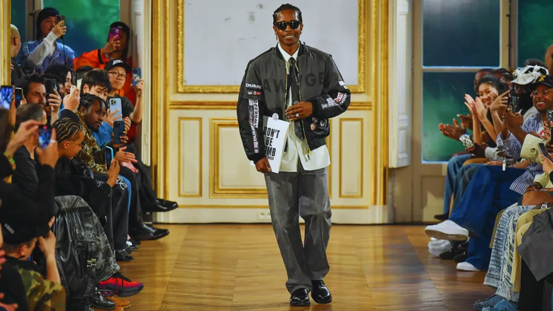 A man wearing sunglasses and a jacket confidently walks down the runway, showcasing a stylish look.