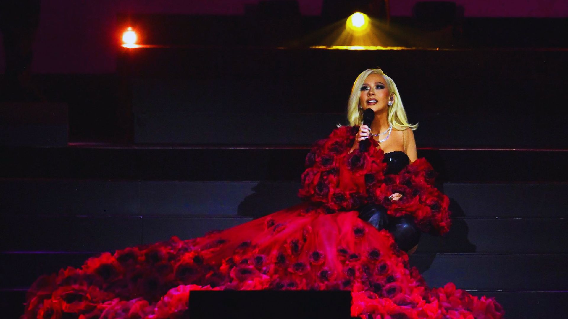 A woman in a red dress stands confidently on stage, engaging the audience with her presence.
y