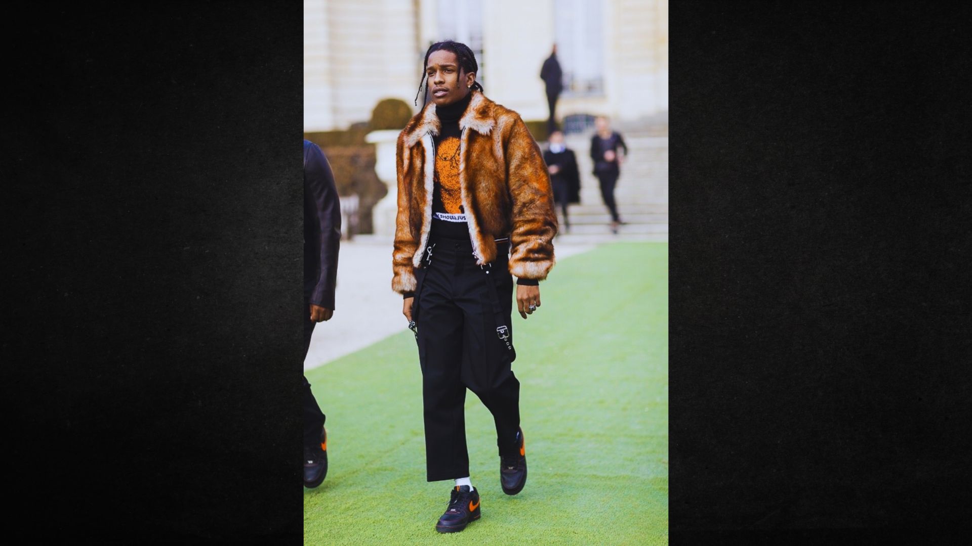 A man walks confidently outdoors, wearing a brown fur jacket and black pants. The setting is urban and stylish, with a relaxed yet fashionable tone.