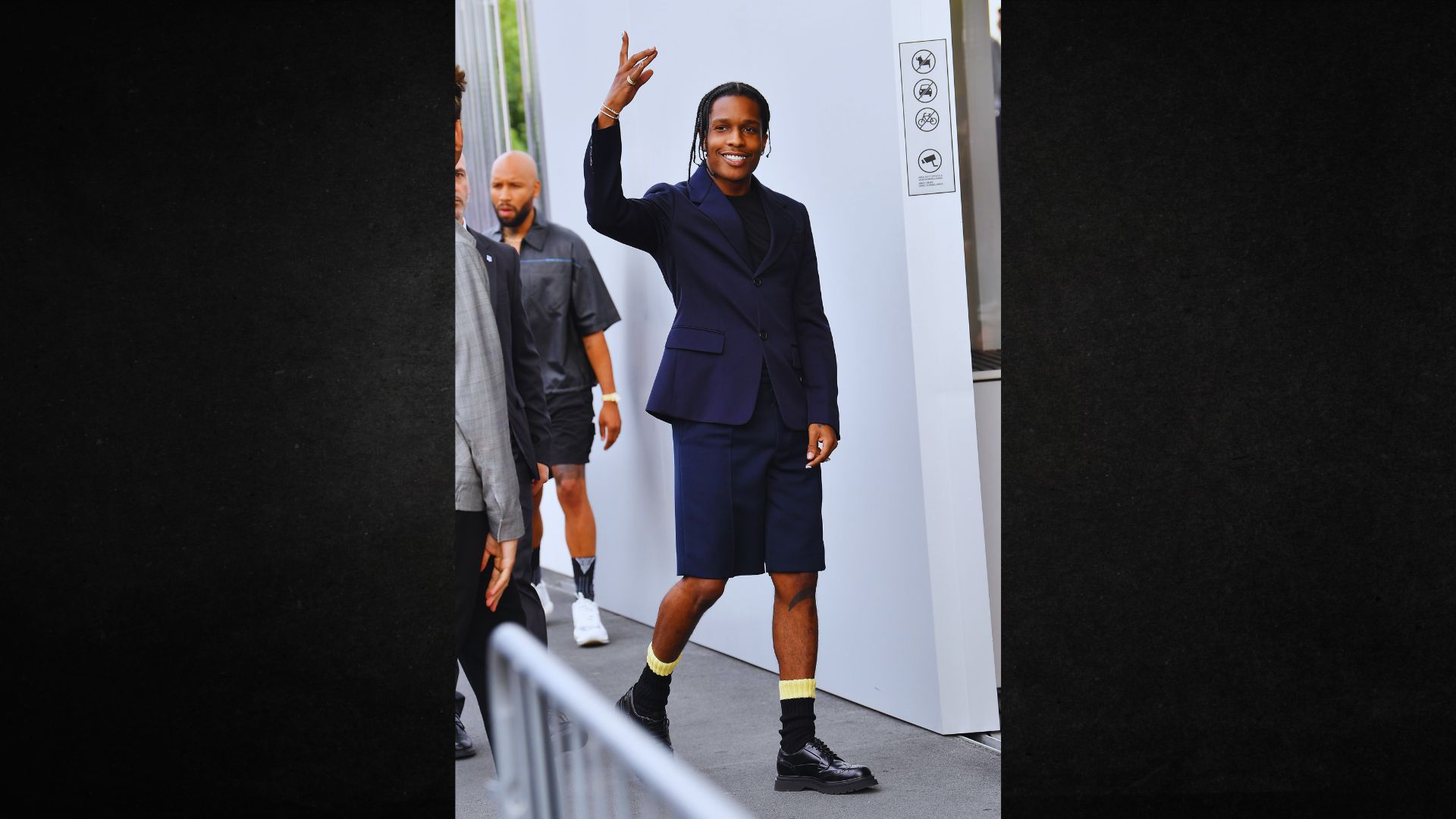 Man in a navy suit and shorts waves with a smile, standing outdoors. He wears yellow socks with black shoes, exuding a stylish, confident vibe.