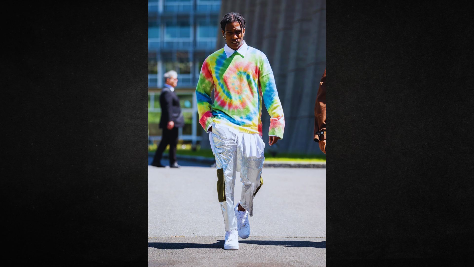 A person walks confidently outside, wearing a vibrant tie-dye sweater and reflective silver pants. The background features modern glass buildings.