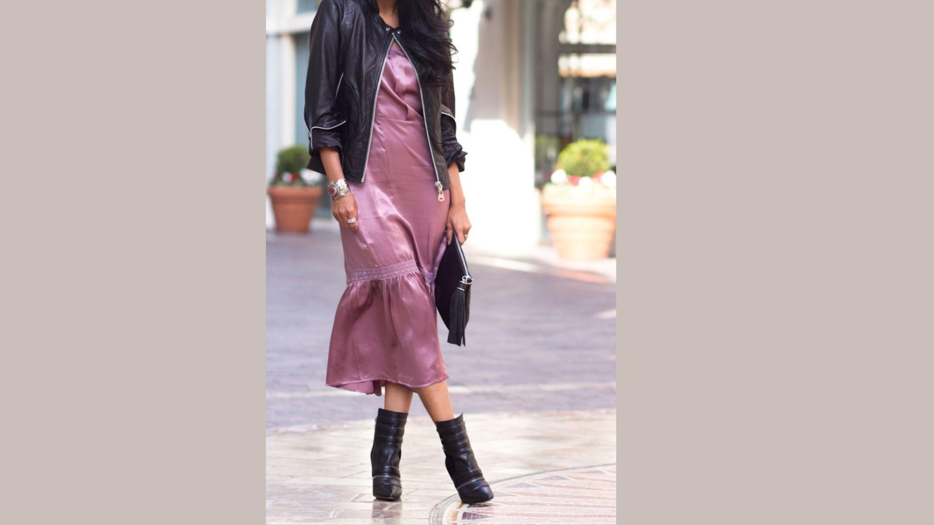 A woman wearing a purple dress and a leather jacket stands confidently, showcasing her stylish outfit.