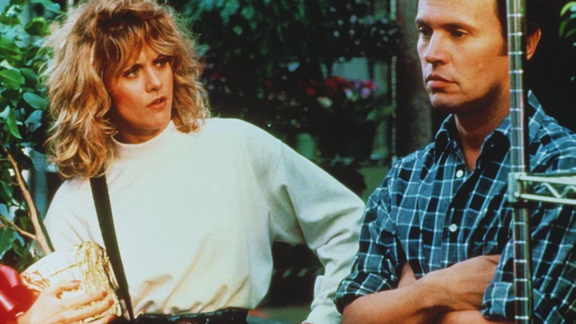 Woman with curly blonde hair in a white sweater looks at a man in a plaid shirt, arms crossed. They stand in a garden center, foliage in the background.