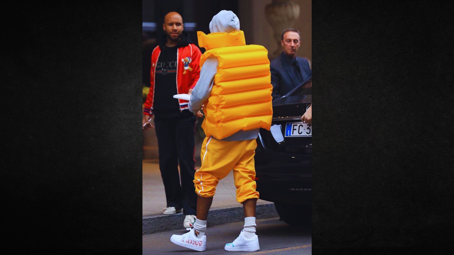 A person wearing an oversized, inflatable orange vest and matching pants stands on a sidewalk. They are near others, creating a whimsical, playful street scene.