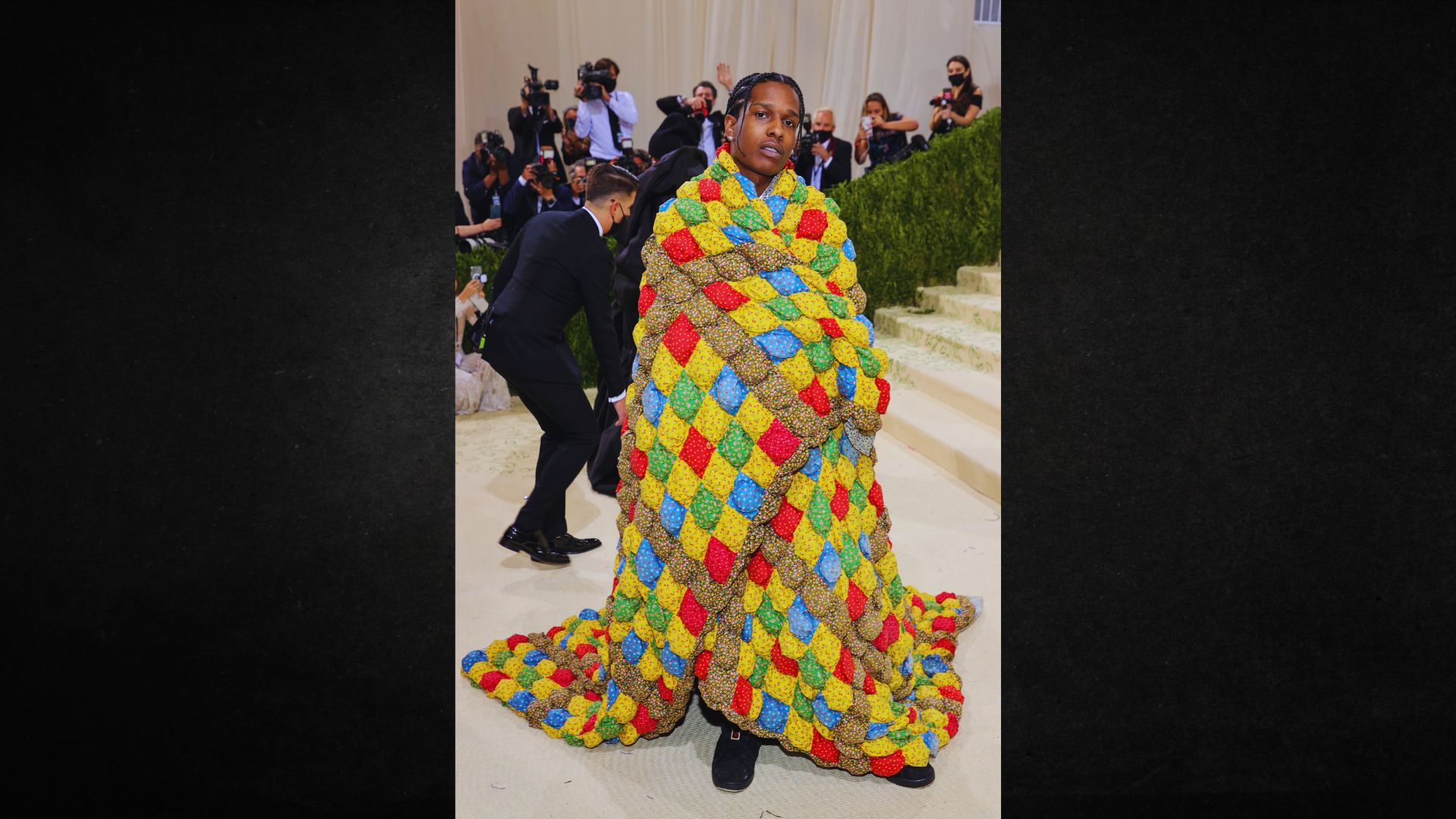 A person is wrapped in a colorful, patterned quilt-like outfit at a formal event, with photographers in the background, evoking a cozy yet fashionable vibe.