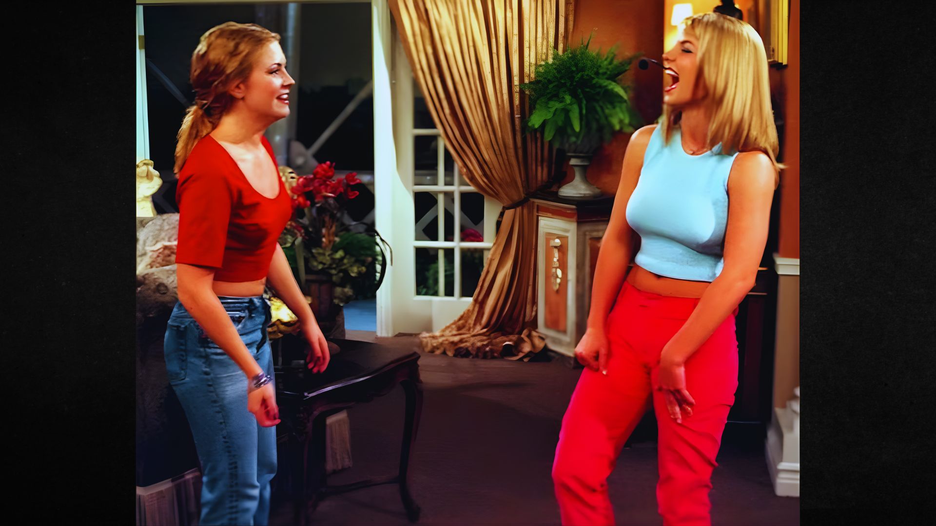 Two women are standing in a living room, laughing and facing each other. One wears a red top and jeans, and the other a blue top with red pants. The background includes drapes, plants, and a decorative cabinet, conveying a cheerful and warm atmosphere.