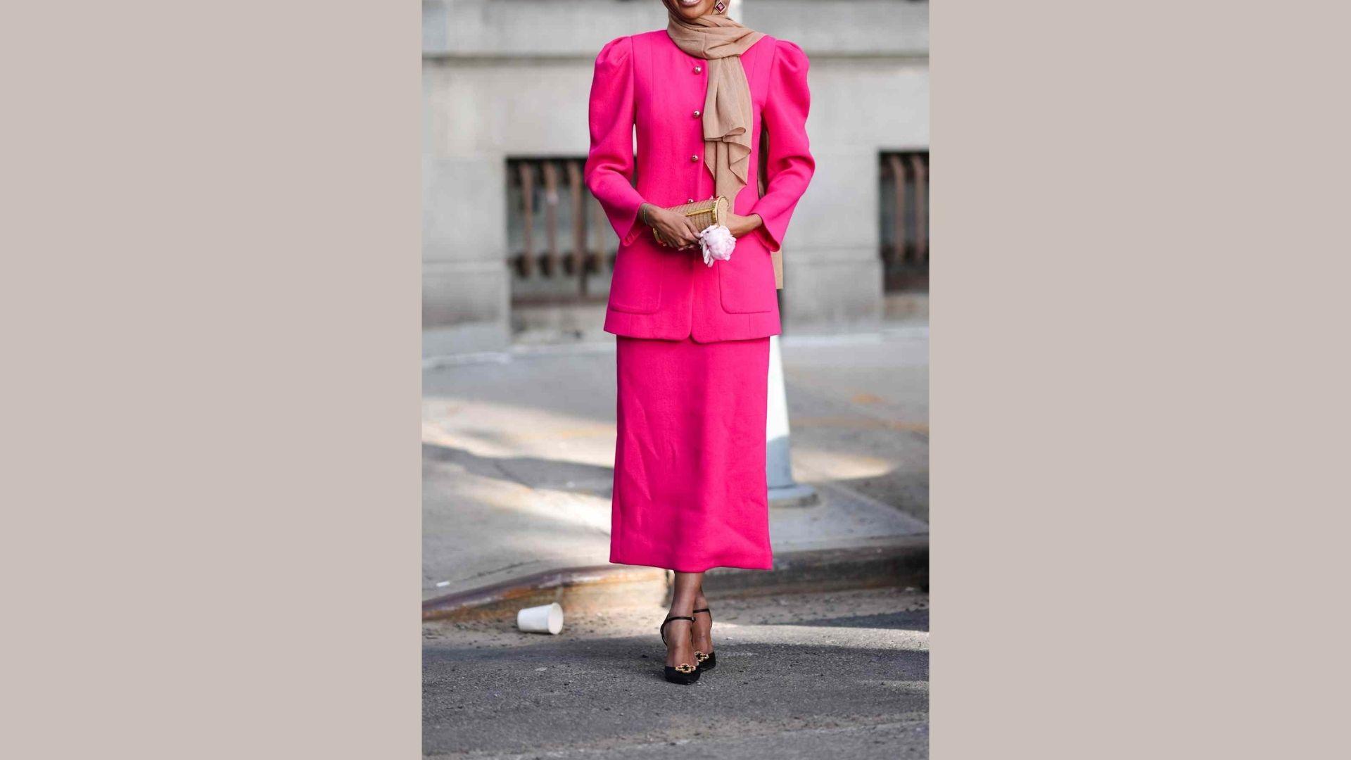 A woman wearing a pink suit and matching scarf stands confidently, showcasing a stylish and professional appearance.