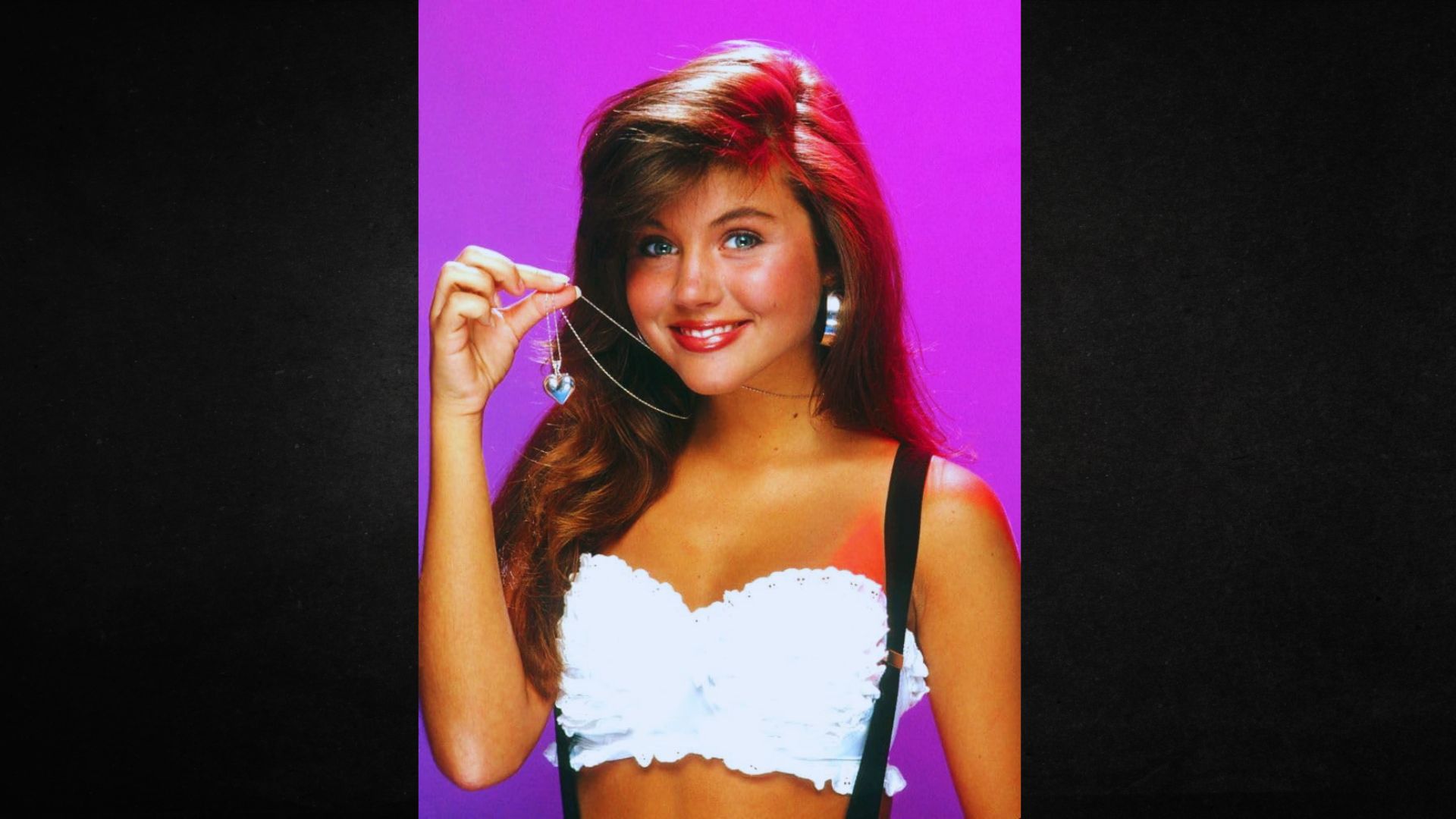 Smiling woman with long hair holds a heart-shaped pendant necklace. She wears a white ruffled crop top and suspenders against a bright pink background.
