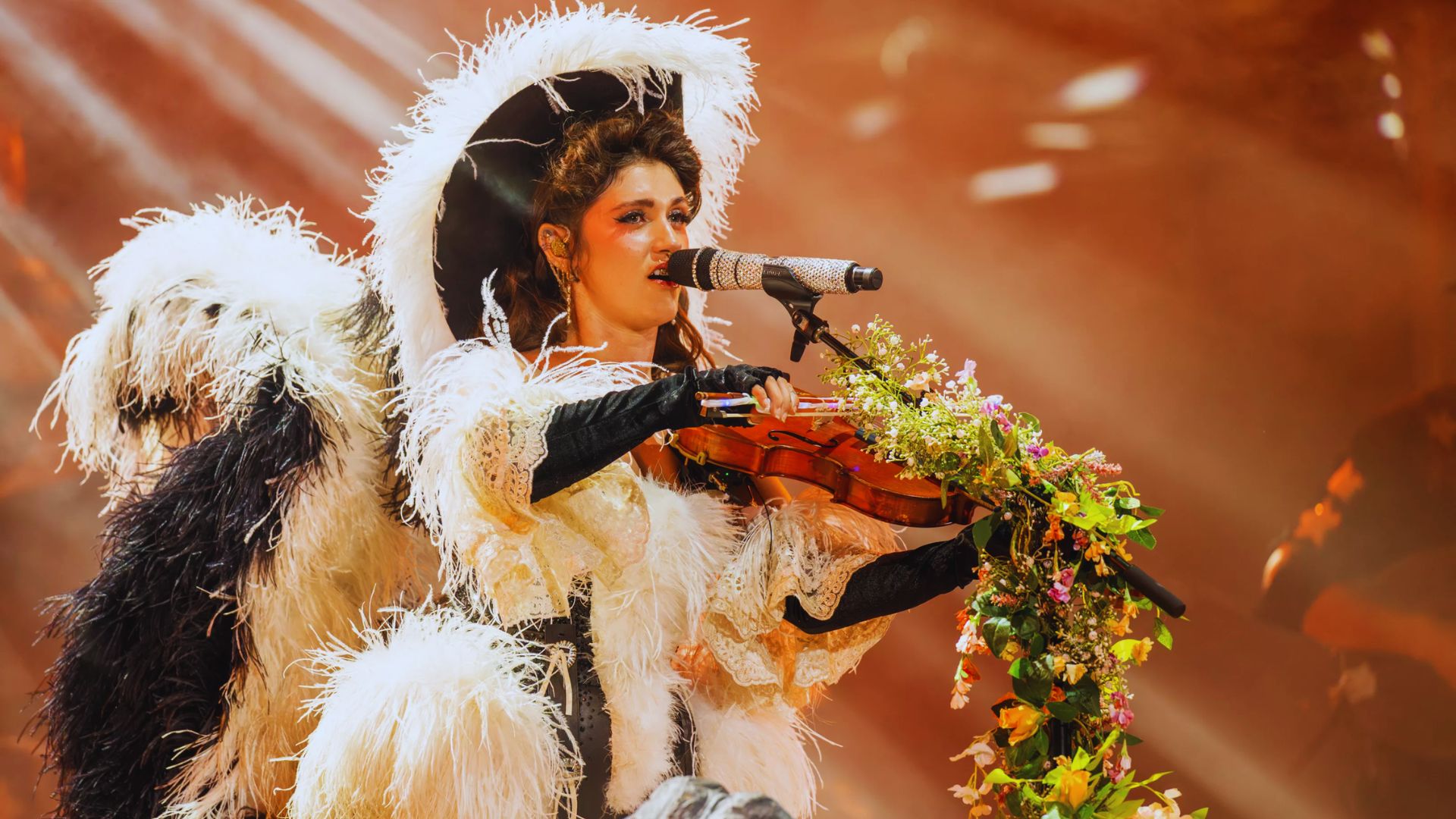 A performer in an elaborate feathered costume plays a violin adorned with flowers. Warm lights create a dramatic, ethereal stage ambiance.