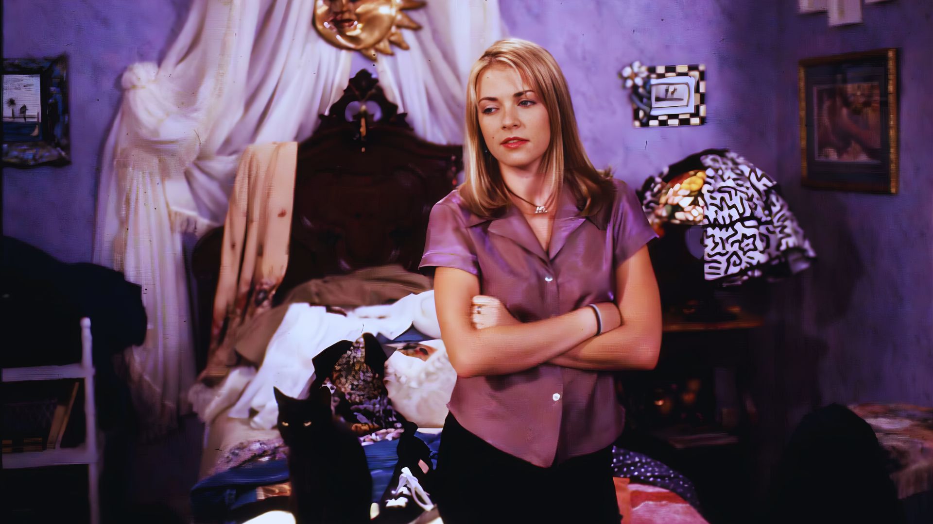 Young woman with long blond hair, wearing a purple blouse, stands with arms crossed in a cluttered bedroom. A black cat is on the bed. The room has purple walls and is decorated with various items, imparting a quirky, cozy feel.