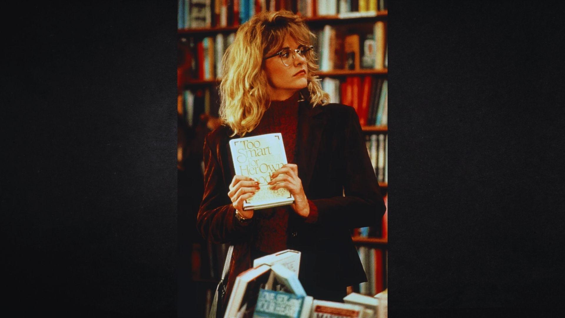 A person with wavy blonde hair and glasses holds a book titled