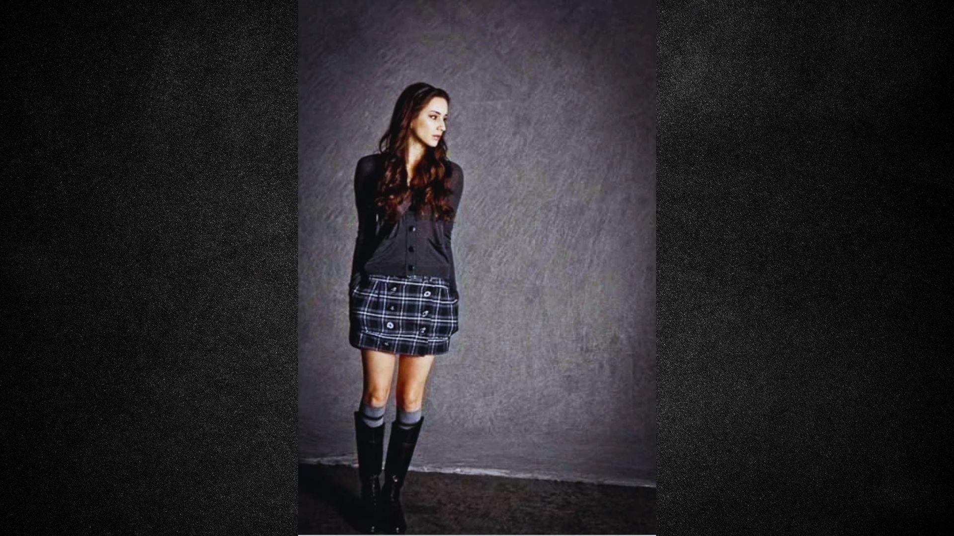 A woman stands confidently for a photo, dressed in a plaid skirt and fashionable boots.