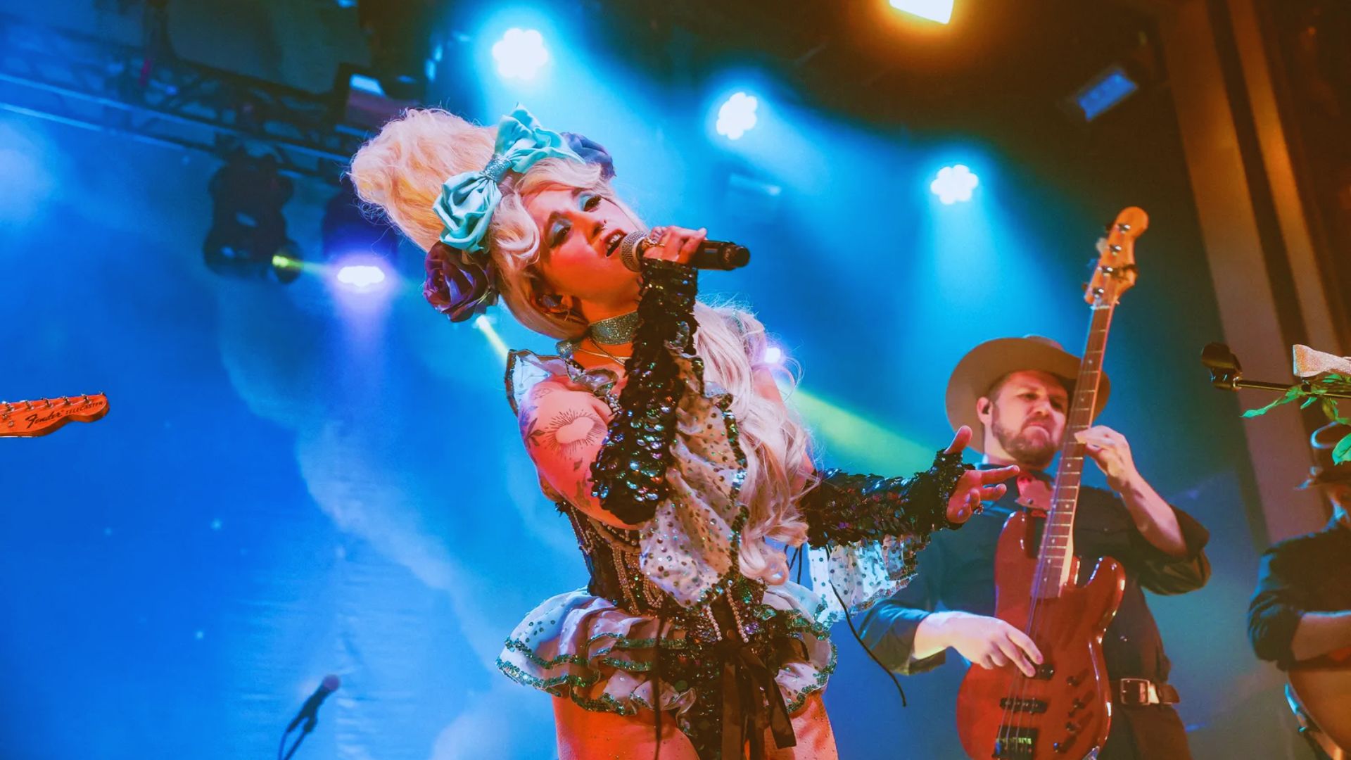 A performer in flamboyant costume, with a large hairpiece and floral details, sings passionately on a colorful stage, surrounded by musicians. Vibrant, dynamic mood.