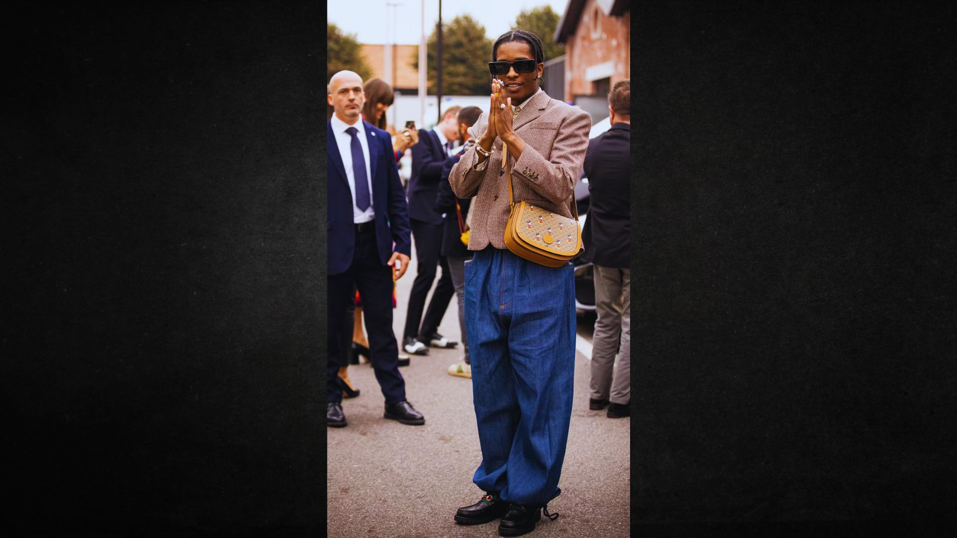 Man in stylish attire stands outdoors, wearing sunglasses, a brown jacket, baggy blue jeans, and a yellow handbag. He appears confident and fashionable.