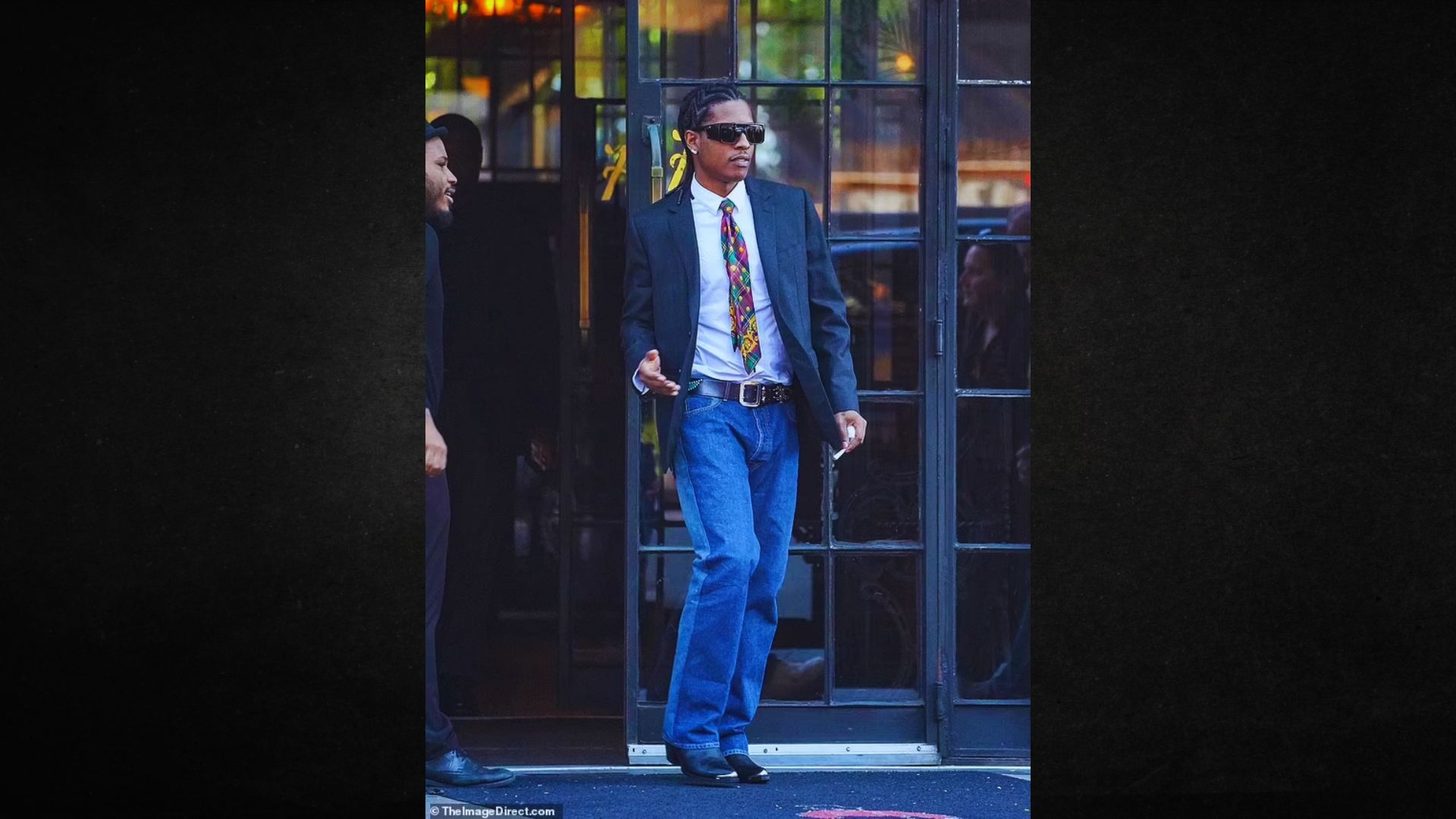 Man wearing sunglasses, a black blazer, and colorful tie exits a building confidently, conveying a relaxed yet stylish vibe.