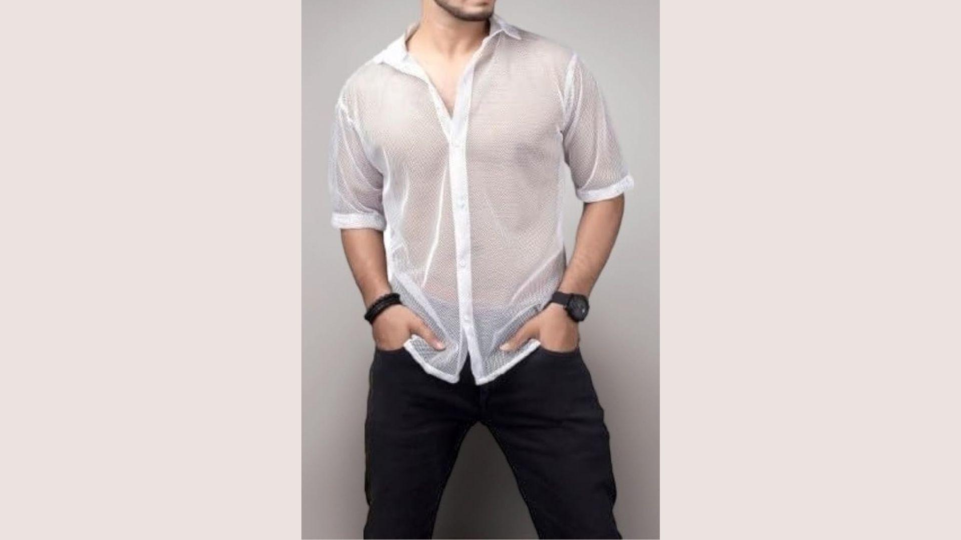 Man in a semi-transparent white shirt and black pants poses confidently with hands in pockets, wearing a black watch, against a gray background.