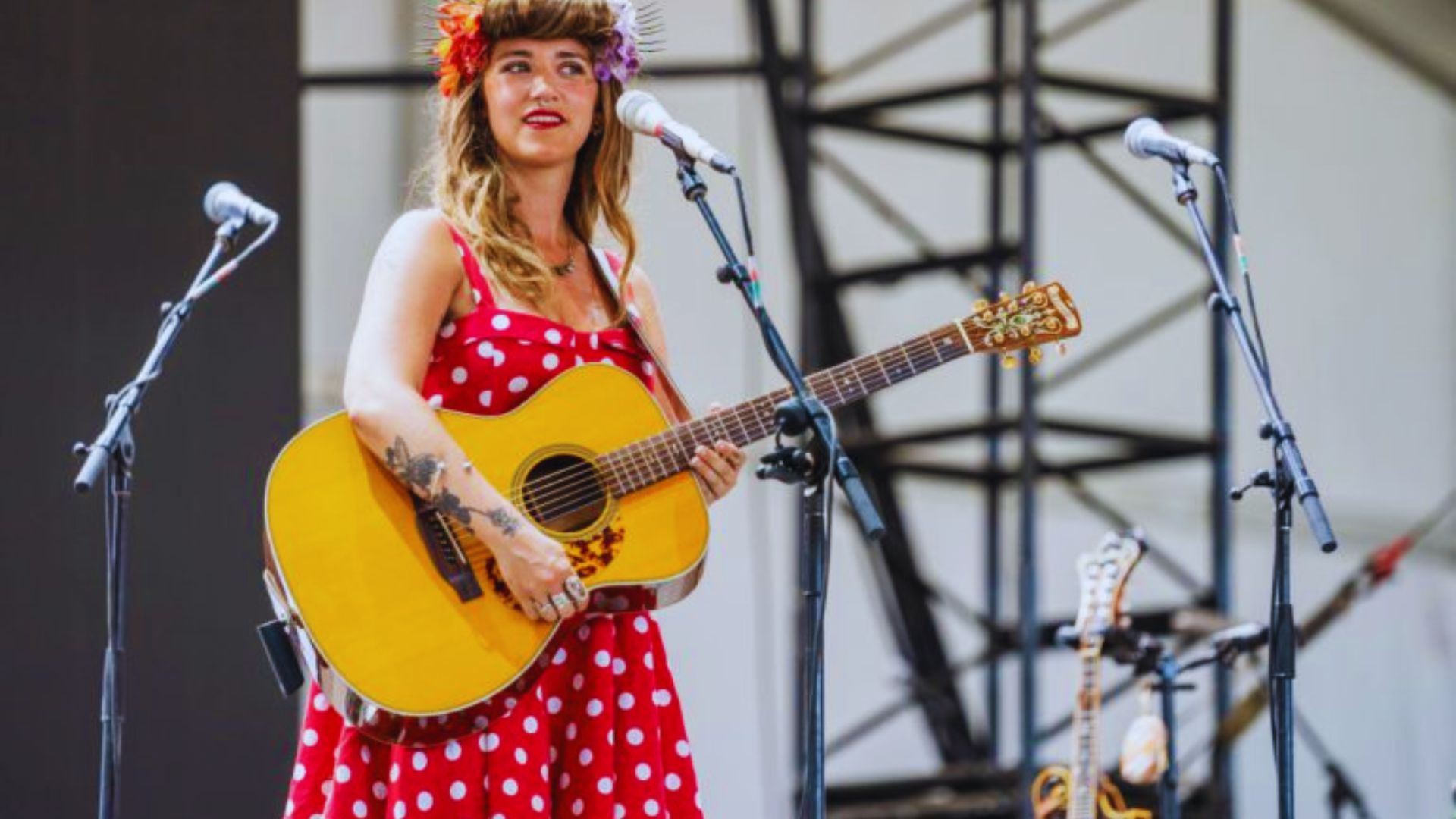 A woman in a red polka dot dress plays a guitar on stage, surrounded by microphones. She has a colorful flower in her hair, conveying an upbeat mood.