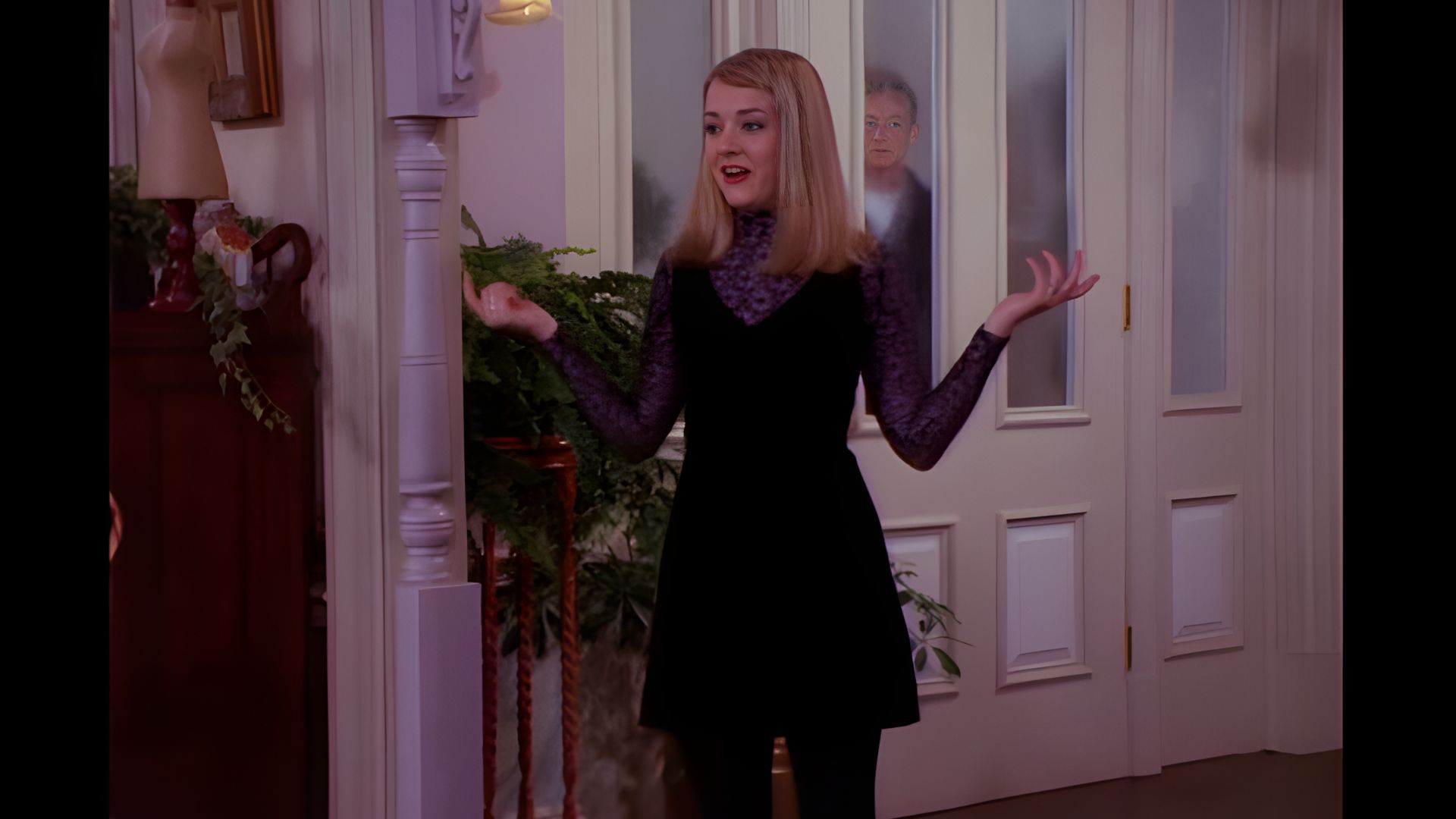 A young woman with blonde hair gestures expressively in a stylish black dress and purple lace top. A person is visible behind her through a glass door.