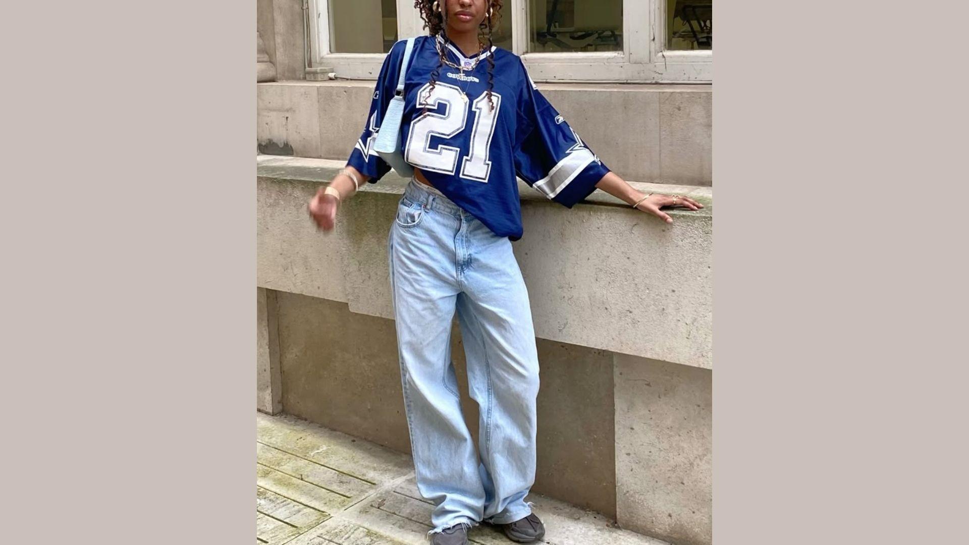 A woman in a jersey and jeans leans casually against a wall, showcasing a relaxed and stylish pose.