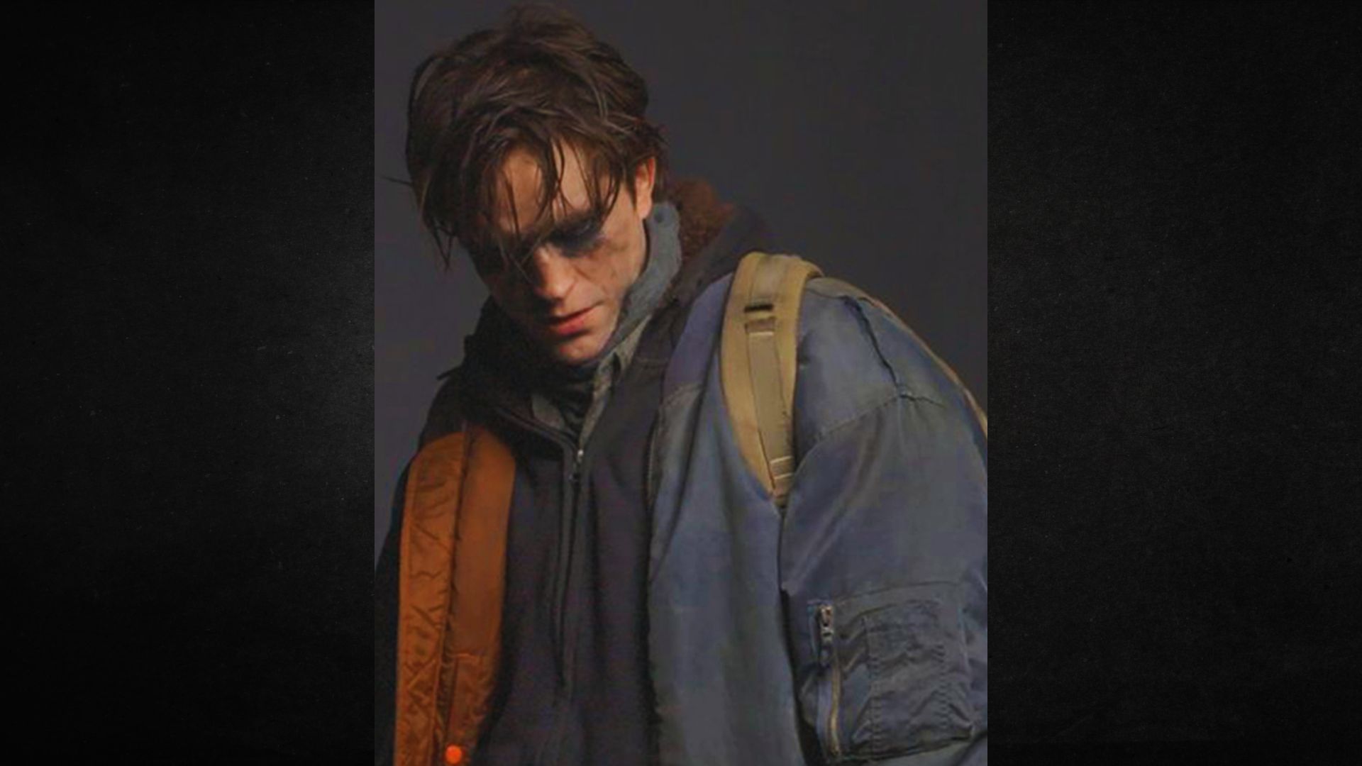 A person with smudged makeup and disheveled hair wears a blue jacket and backpack, looking down with a somber expression against a dark background.
