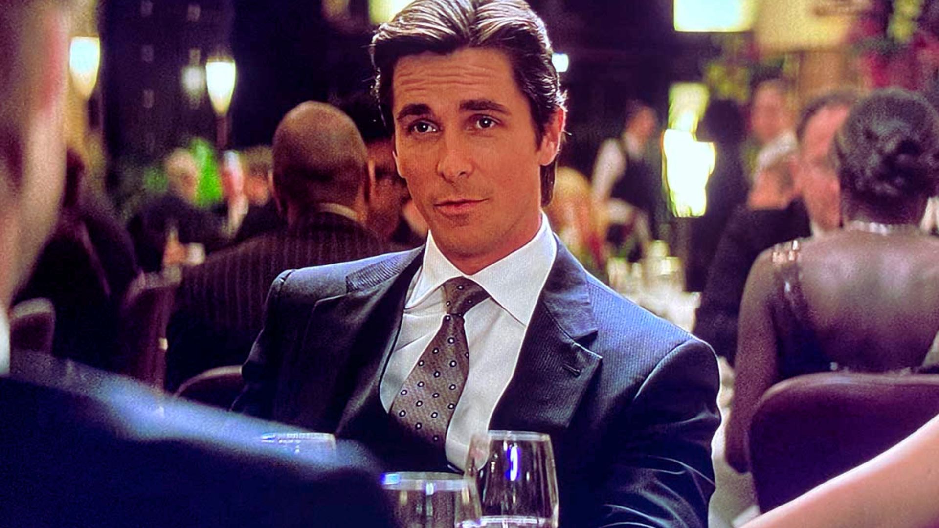 A man in a suit and tie sits at a formal dinner table, exuding confidence. Soft lighting and blurred background suggest an elegant, upscale setting.