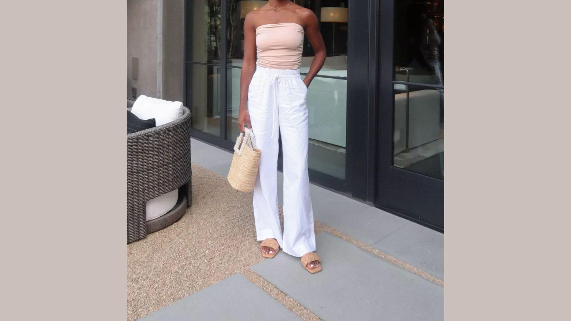 A woman wearing white pants and a pink top stands confidently, showcasing a casual yet stylish outfit.