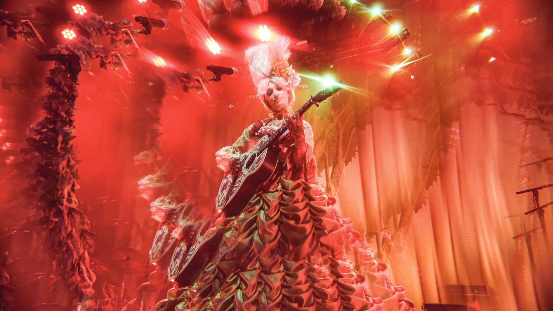 A performer in an elaborate, ruffled costume with a high wig plays guitar under vibrant red lighting, creating a surreal, theatrical atmosphere.