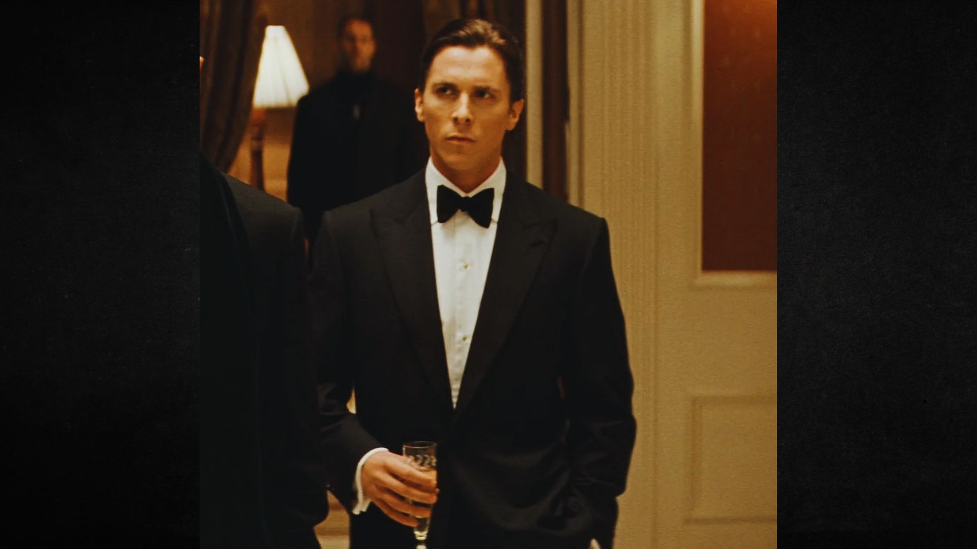 A man in a tuxedo stands at a formal event, holding a drink. He appears focused and calm. Soft lighting and elegant decor create a sophisticated atmosphere.