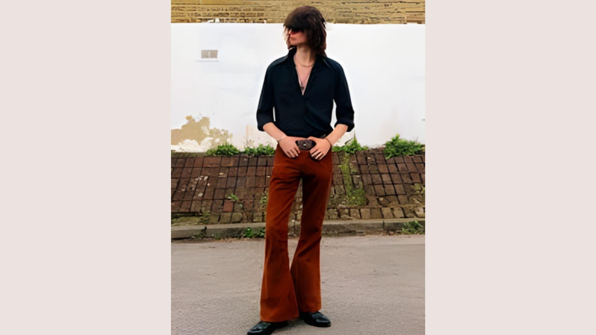 A person stands confidently outdoors wearing a black shirt and brown flared pants, conveying a retro fashion vibe. The background features a white wall and greenery.