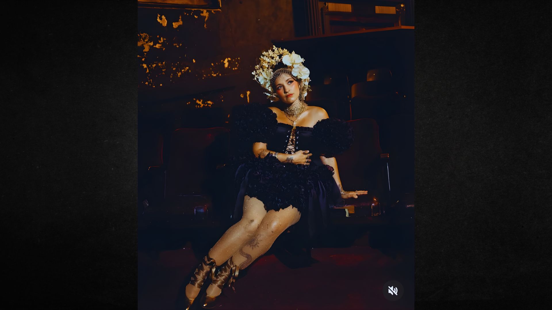 A woman in an elegant black dress with puffed sleeves sits in a dimly lit theater. She wears a white floral headdress, exuding a regal and ethereal vibe.