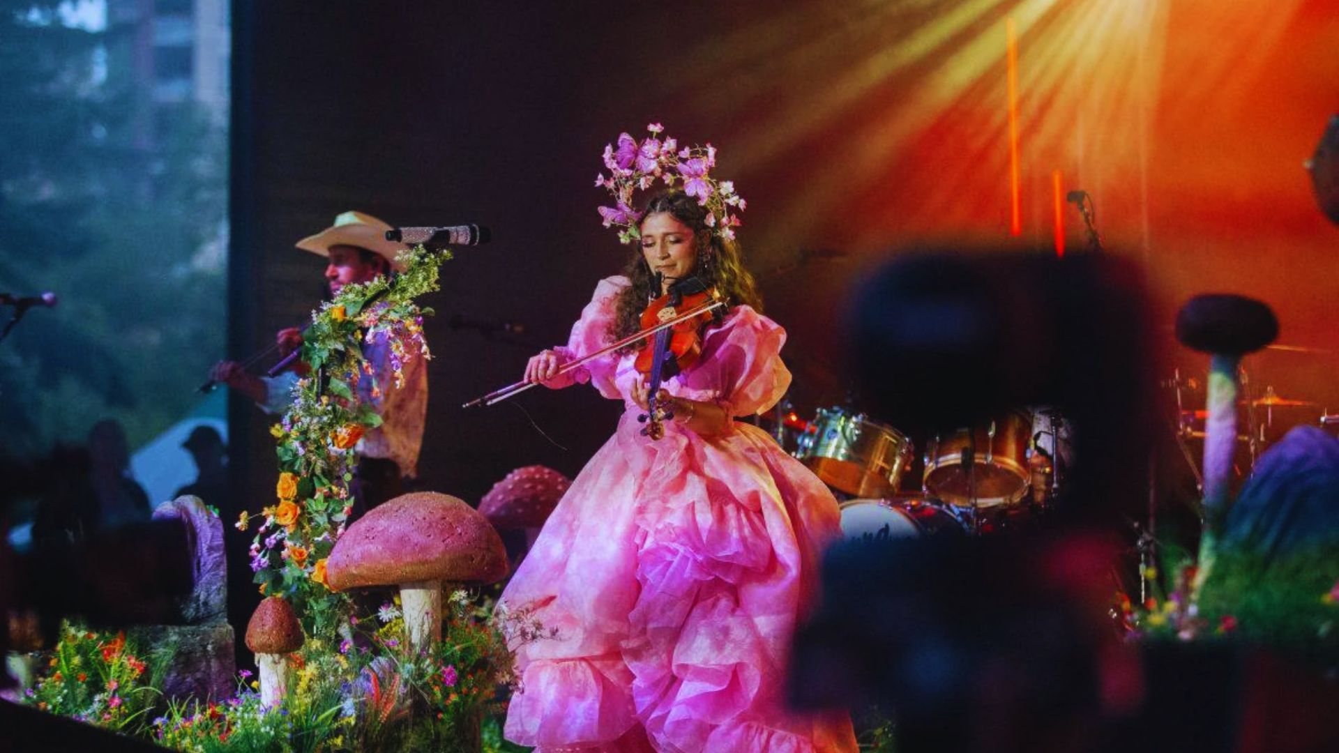 A performer in a pink dress and floral headpiece plays violin on stage amidst colorful flowers and mushroom props, creating a whimsical, enchanting atmosphere.